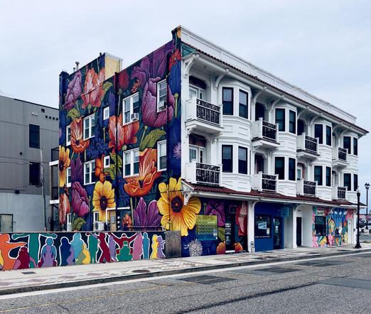 A color, wide-angle photograph that captures a three-story corner building in Atlantic City on an overcast winter afternoon. The building features an expansive, vibrant mural of oversized flowers covering its left side and wrapping around the ground floor. The mural is a dense composition of tulips and sunflowers in deep shades of purple, orange, yellow, and blue, creating a striking contrast against the building’s upper white facade. To the far left, a low wall continues the artistic theme with a series of stylized, colorful silhouettes of people in various poses. At the street level, small storefronts are integrated into the mural, including one with a blue sign for "Hayday Coffee" and a cannabis dispensary with whimsical, cartoonish illustrations. The architectural style of the building combines traditional elements with modern artistic flair. The upper two floors on the right are painted a crisp white, featuring classic bay windows and small balconies with ornate black wrought-iron railings. A terracotta-style tiled roofline runs above the ground-floor shops, separating the residential-looking upper levels from the commercial street front. The photo is taken from across an asphalt road, capturing the building from an angled perspective that highlights the transition from the bold, floral street art on the left to the more structured, white-paneled architecture on the right.