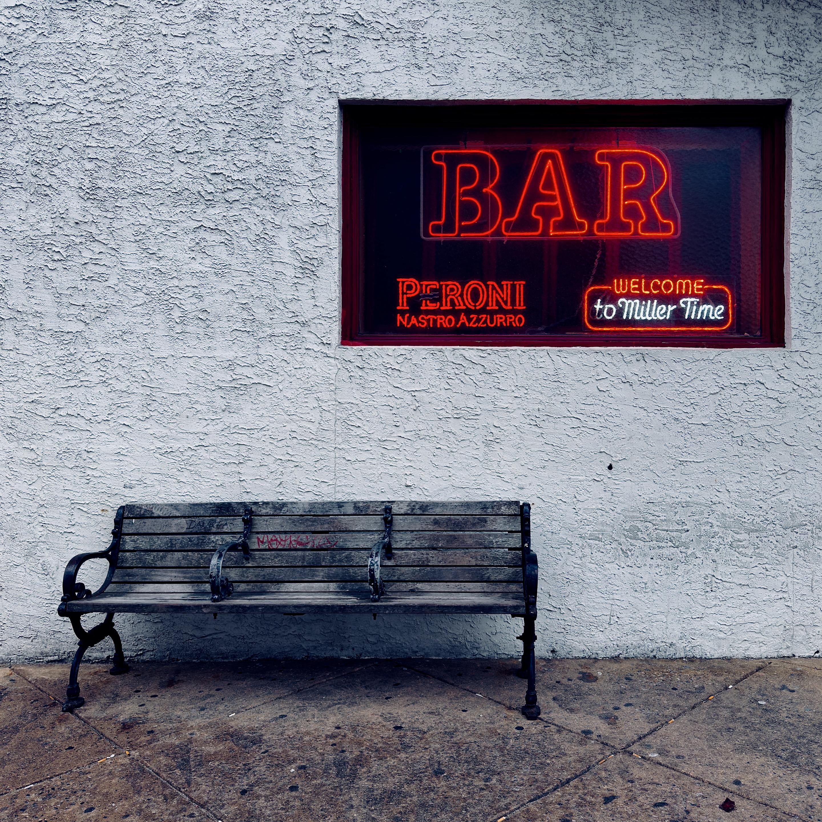 A color photograph in square aspect ratio that captures the street scene along Iowa Avenue in Atlantic City. The image features a weathered, empty wooden bench sitting against a stark, light-colored stucco wall outside of Tony’s Baltimore Grill. The bench shows signs of age with worn slats and some red graffiti scrawled across the middle section. Above the bench, a dark window frame houses several glowing neon signs. The largest sign in the center displays the word "BAR" in bold, red-orange neon outlines. Below and to the left, smaller neon lettering advertises "PERONI NASTRO AZZURRO," while a rectangular sign to the right features a "WELCOME to Miller Time" graphic with a red border. The lighting in the photo portrays the gray, overcast day. The ground is a paved sidewalk with a diagonal grid pattern, dotted with a few small pieces of debris. The overall mood is quiet and somewhat desolate.