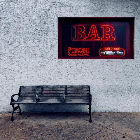 A color photograph in square aspect ratio that captures the street scene along Iowa Avenue in Atlantic City. The image features a weathered, empty wooden bench sitting against a stark, light-colored stucco wall outside of Tony’s Baltimore Grill. The bench shows signs of age with worn slats and some red graffiti scrawled across the middle section. Above the bench, a dark window frame houses several glowing neon signs. The largest sign in the center displays the word "BAR" in bold, red-orange neon outlines. Below and to the left, smaller neon lettering advertises "PERONI NASTRO AZZURRO," while a rectangular sign to the right features a "WELCOME to Miller Time" graphic with a red border. The lighting in the photo portrays the gray, overcast day. The ground is a paved sidewalk with a diagonal grid pattern, dotted with a few small pieces of debris. The overall mood is quiet and somewhat desolate.