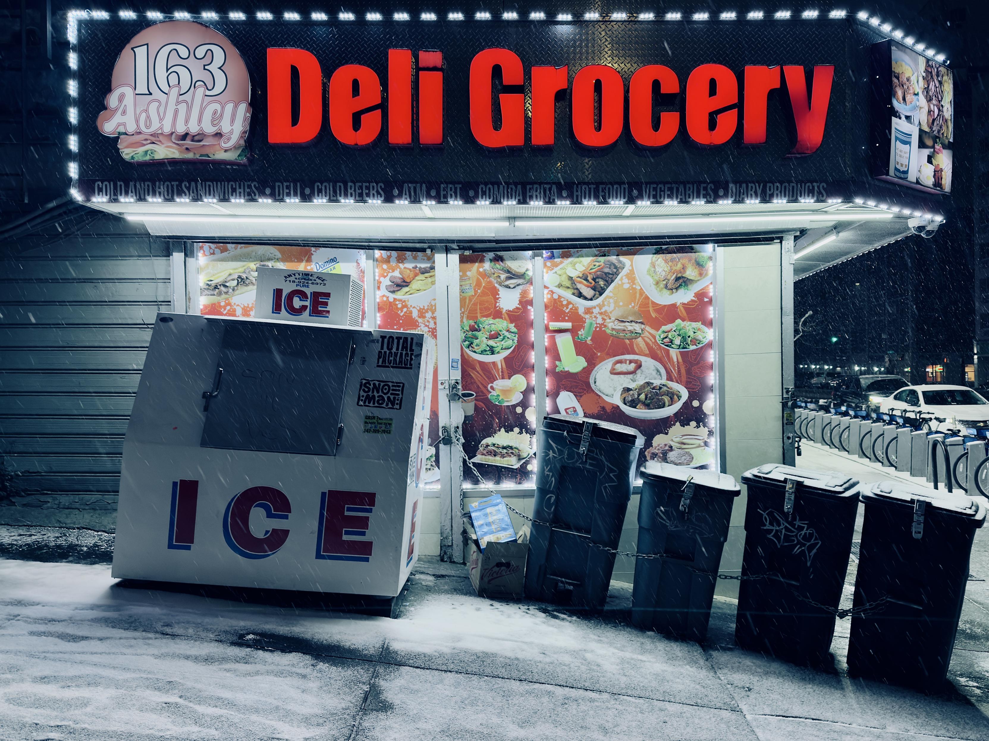 A color, medium-angle photograph that captures the exterior of the "163 Ashley Deli Grocery" corner store in the Bronx during a nighttime snowfall. The storefront is dominated by a large black sign with the words "Deli Grocery" glowing in bold, red neon-style lettering. To the left of the text, a circular logo features a sandwich and the numbers "163” and word “Ashley.” Below the main sign, a thin backlit strip lists various offerings such as "Cold and Hot Sandwiches," "Cold Beers," “ATM,” and "Hot Food." The large glass windows are covered with vibrant decals showcasing photos of various dishes, including salads, rice plates, and sandwiches, which are illuminated by strings of white fairy lights bordering the window frames. In the foreground, the steep inclined sidewalk is covered in a thin, fresh layer of white snow, and visible white streaks of active snowfall. To the left stands a large, white "ICE" refrigeration box with the word "ICE" printed in large red and blue block letters. Next to it, a row of four plastic trash bins are chained together and to the storefront for security. To the far right, a Citi Bike docking station is visible, with a row of bikes parked and a white car driving past in the distance. The overall atmosphere is cold and dramatic in tone.