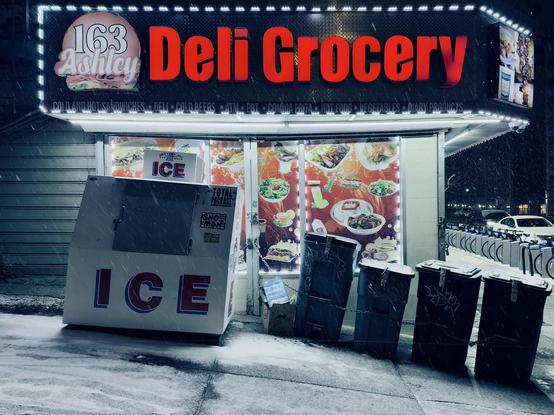 A color, medium-angle photograph that captures the exterior of the "163 Ashley Deli Grocery" corner store in the Bronx during a nighttime snowfall. The storefront is dominated by a large black sign with the words "Deli Grocery" glowing in bold, red neon-style lettering. To the left of the text, a circular logo features a sandwich and the numbers "163” and word “Ashley.” Below the main sign, a thin backlit strip lists various offerings such as "Cold and Hot Sandwiches," "Cold Beers," “ATM,” and "Hot Food." The large glass windows are covered with vibrant decals showcasing photos of various dishes, including salads, rice plates, and sandwiches, which are illuminated by strings of white fairy lights bordering the window frames. In the foreground, the steep inclined sidewalk is covered in a thin, fresh layer of white snow, and visible white streaks of active snowfall. To the left stands a large, white "ICE" refrigeration box with the word "ICE" printed in large red and blue block letters. Next to it, a row of four plastic trash bins are chained together and to the storefront for security. To the far right, a Citi Bike docking station is visible, with a row of bikes parked and a white car driving past in the distance. The overall atmosphere is cold and dramatic in tone.