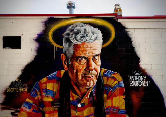 A color, medium-angle photograph of a vibrant memorial mural of Anthony Bourdain painted on an exterior white brick wall of Tony’s Baltimore Grill in Atlantic City. The portrait features Bourdain from the chest up, rendered in a bold, expressive style with visible, blocky brushstrokes and a warm color palette of oranges, yellows, and reds. He wears a multi-colored plaid shirt in shades of red, blue, and yellow. His silver hair is meticulously detailed with swirling white and gray highlights, and a glowing yellow halo is spray-painted behind his head. The background immediately behind him is a dark, shadowy silhouette that makes the portrait pop against the lighter brick. To the right of the portrait, the name "ANTHONY BOURDAIN" is written in white, stylized capital letters, topped with a small, minimalist halo icon. On the far left, the artist’s handle, "@SCOTTIE.MARSH," is tagged in yellow script. The wall has a red trim along the roofline and metal exhaust pipes visible above. The sky is a light grey, on an overcast afternoon.