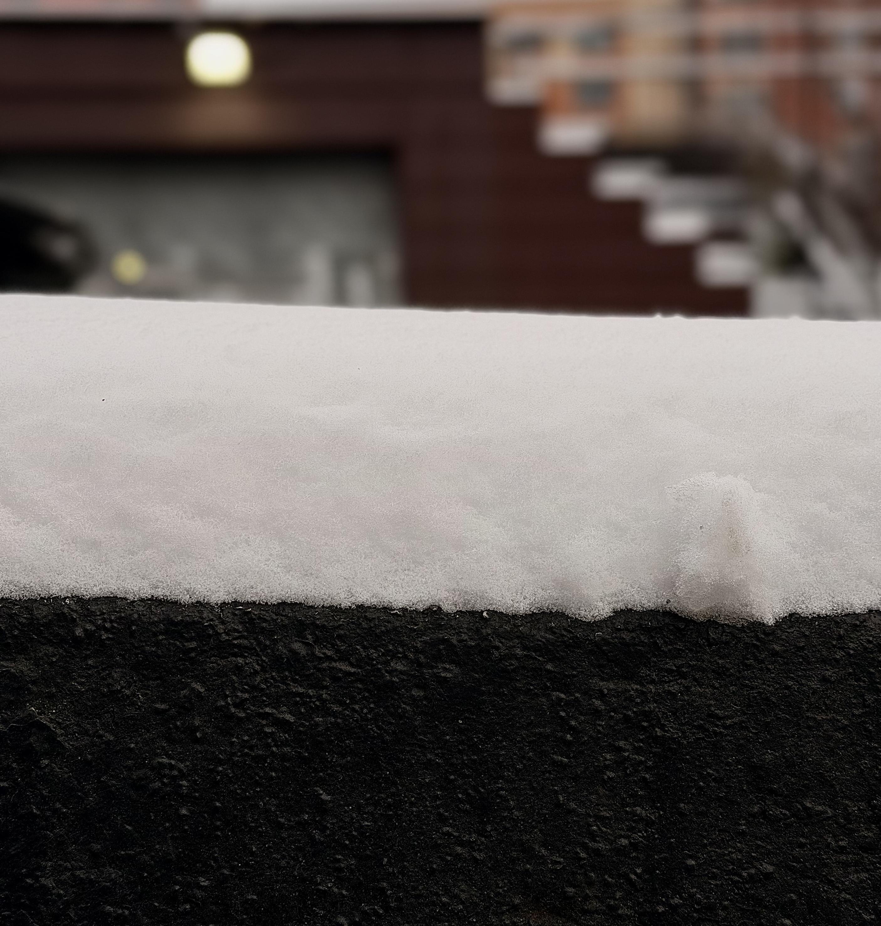 A color, close-up photograph that captures a fresh layer of snow resting atop a dark, textured stone wall. The snow is clean and white, showing a granular, crystalline texture of a light snowfall. It forms a soft, even blanket about three inches thick, with a small, jagged clump of snow protruding slightly on the right side from running my finger across it, breaking the otherwise smooth horizontal line. The dark base beneath provides a sharp tonal contrast. The background is softly blurred with a shallow depth of field, creating a bokeh effect that emphasizes the crisp details of the snow in the foreground. In the distance, the warm brown tones of a building and the faint, out-of-focus shapes of a staircase are visible. A single, glowing yellow light orb is situated on the building above a rolling gate. The tone is muted from the overcast morning.