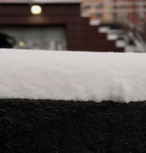 A color, close-up photograph that captures a fresh layer of snow resting atop a dark, textured stone wall. The snow is clean and white, showing a granular, crystalline texture of a light snowfall. It forms a soft, even blanket about three inches thick, with a small, jagged clump of snow protruding slightly on the right side from running my finger across it, breaking the otherwise smooth horizontal line. The dark base beneath provides a sharp tonal contrast. The background is softly blurred with a shallow depth of field, creating a bokeh effect that emphasizes the crisp details of the snow in the foreground. In the distance, the warm brown tones of a building and the faint, out-of-focus shapes of a staircase are visible. A single, glowing yellow light orb is situated on the building above a rolling gate. The tone is muted from the overcast morning.
