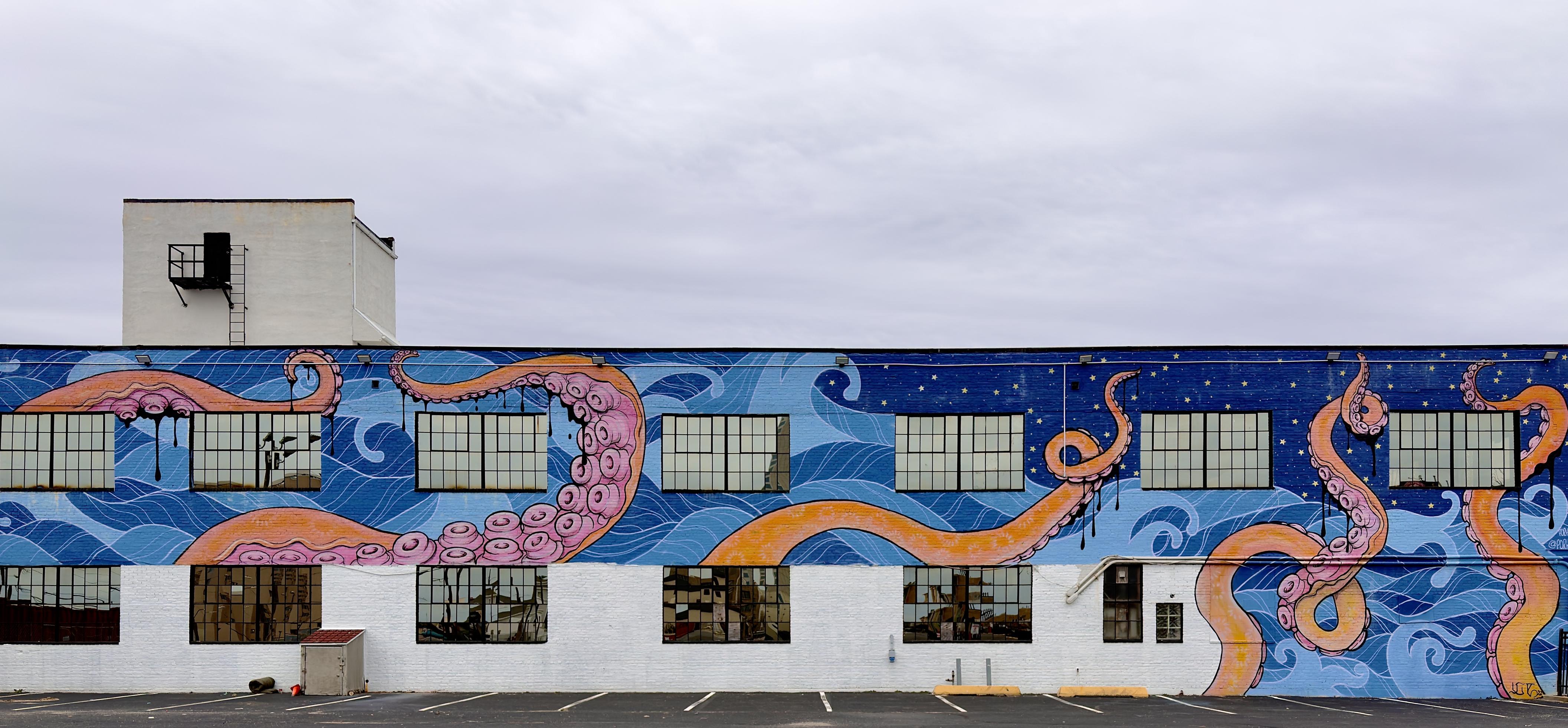 A color, wide-angle photograph in landscape orientation that captures a massive, vibrant mural painted across a long, two-story industrial building under a pale, overcast winter afternoon sky in Atlantic City. The mural features an enormous octopus with thick, swirling tentacles in shades of coral pink and golden orange, textured with detailed suction cups. The creature is set against a backdrop of stylized ocean waves in various shades of blue and periwinkle, which transition on the right side of the building into a dark indigo night sky dotted with small, yellow stars. Black, ink-like drips trail down from the tentacles and window frames, adding a sense of movement and depth to the artwork. The building itself is white with two rows of large, grid-style industrial windows that are integrated into the art; the upper windows sit within the blue waves, while the lower windows are set against the plain white wall of the first floor. A small white penthouse structure with a black metal ladder and door sits atop the roof on the left side. In the foreground, an empty asphalt parking lot shows yellow painted stall lines stretching toward the base of the building.