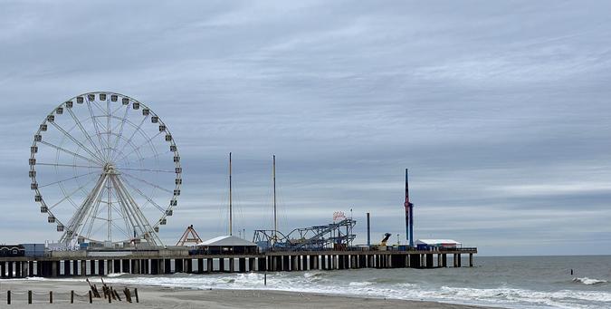 A color, wide-angle photograph that captures the Steel Pier in Atlantic City, New Jersey, extending into the ocean under a heavy, overcast gray sky. The pier is supported by a long row of dark vertical pilings that disappear into the churning, foamy white surf of the Atlantic Ocean. On the left side of the pier, a massive Ferris wheel dominates the skyline, its circular frame fitted with numerous passenger gondolas and accented by subtle rainbow changing lighting. To the right of the wheel, various amusement park attractions are visible, including several tall, thin vertical drop towers, a blue-railed roller coaster, and small white structures for concession stands. An illuminated silver sign on the far left reads “STEEL PIER.” In the foreground, a wide stretch of light gray sandy beach occupies the lower part of the frame. The shoreline is scattered with small white seagulls near the water's edge. On the far left, a low wooden fence with dark posts and rope is partially buried in the sand, while several weathered wooden beams stick out of the beach at an angle. The water is a dark, murky gray-green, with waves breaking into white foam as they roll toward the shore. The horizon line is flat and distant, meeting the thick layer of clouds that creates a moody scene.