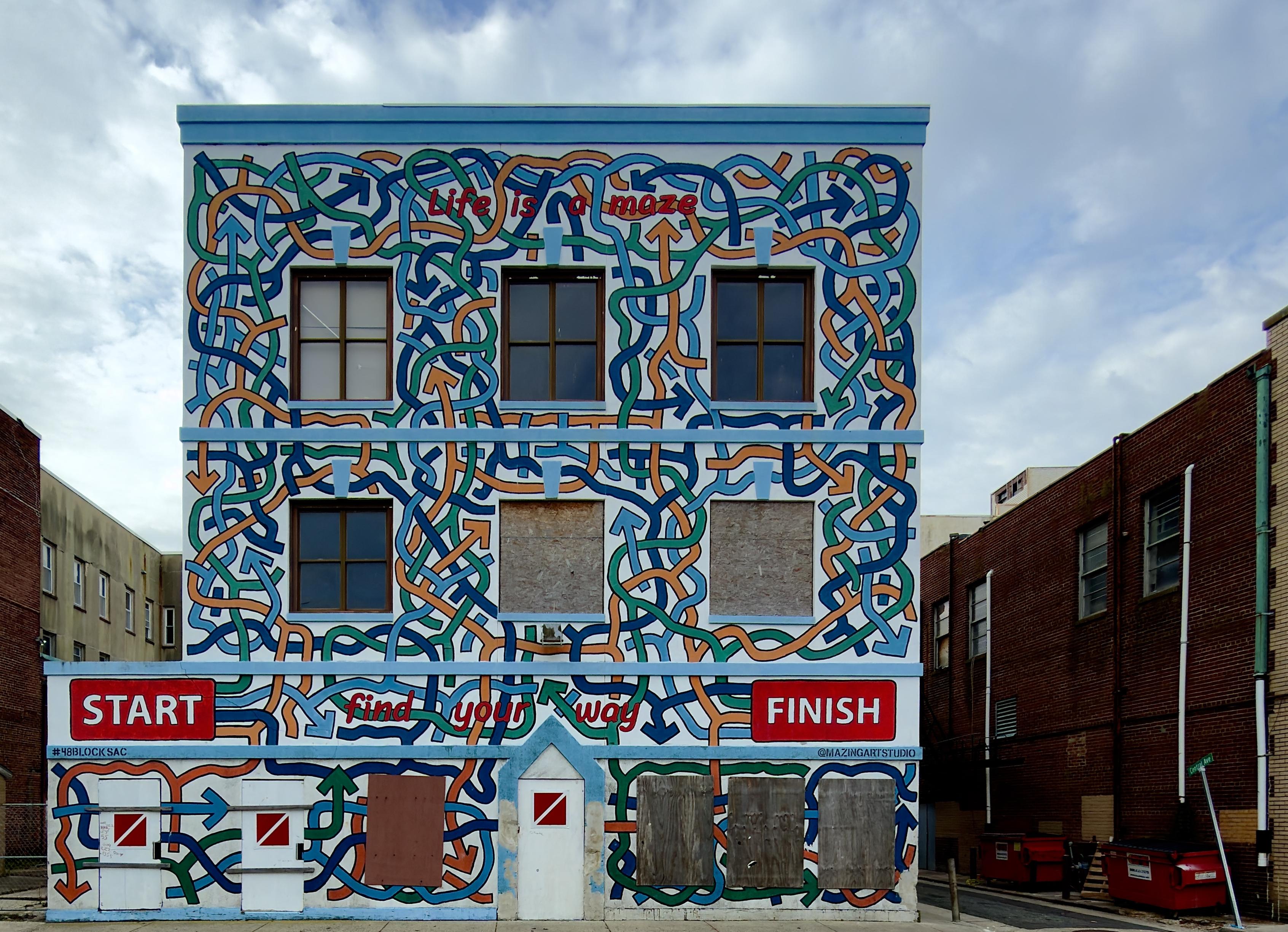 A color, medium-angle photograph that captures a vibrant, three-story building in Atlantic City that is completely covered in a mural that depicts a complex, interlocking maze of colorful lines. The mural features thick, intertwined paths in shades of teal, navy blue, and orange set against a white background, with many paths ending in arrowheads pointing in various directions. The architectural elements of the building, including six rectangular windows on the upper floors and several doors at ground level, are integrated into the design. Some windows are clear, while others are boarded up with plywood, and the ground-level doors are painted white with small red and white warning symbols. The mural includes several pieces of text that emphasize its theme. Across the top, the phrase "Life is a maze" is written in red, lowercase letters. Near the bottom, a large red rectangular sign on the left reads "START" in bold white capital letters, while a matching sign on the right reads "FINISH." Between these two signs, the phrase "find your way" is scripted in red lowercase letters across the maze paths. In the bottom left corner, the hashtag #48BLOCKSAC is visible, and on the bottom right, the artist's handle @MAZINGARTSTUDIO is credited. The building is situated on a city street under a cloudy winter morning sky, flanked by a plain beige building on the left and a red brick building with an alleyway and red dumpsters on the right.