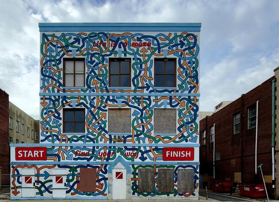 A color, medium-angle photograph that captures a vibrant, three-story building in Atlantic City that is completely covered in a mural that depicts a complex, interlocking maze of colorful lines. The mural features thick, intertwined paths in shades of teal, navy blue, and orange set against a white background, with many paths ending in arrowheads pointing in various directions. The architectural elements of the building, including six rectangular windows on the upper floors and several doors at ground level, are integrated into the design. Some windows are clear, while others are boarded up with plywood, and the ground-level doors are painted white with small red and white warning symbols. The mural includes several pieces of text that emphasize its theme. Across the top, the phrase "Life is a maze" is written in red, lowercase letters. Near the bottom, a large red rectangular sign on the left reads "START" in bold white capital letters, while a matching sign on the right reads "FINISH." Between these two signs, the phrase "find your way" is scripted in red lowercase letters across the maze paths. In the bottom left corner, the hashtag #48BLOCKSAC is visible, and on the bottom right, the artist's handle @MAZINGARTSTUDIO is credited. The building is situated on a city street under a cloudy winter morning sky, flanked by a plain beige building on the left and a red brick building with an alleyway and red dumpsters on the right.