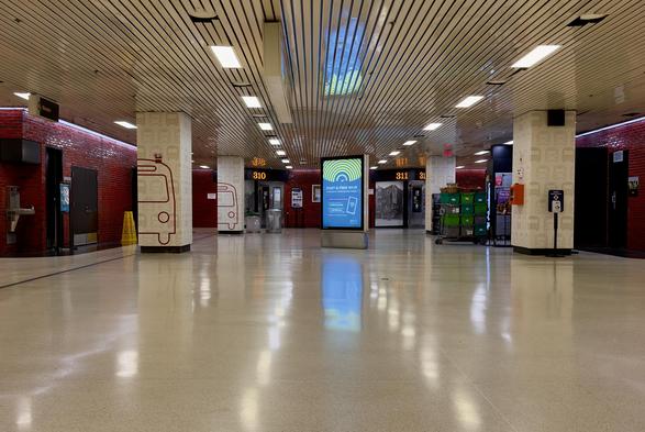 A color, wide-angle photograph that captures a wide, desolate interior view of the Port Authority Bus Terminal in New York City, very early on Christmas Day morning, characterized by its distinctive mid-century modern aesthetic. The room is dominated by high-gloss, cream-colored floors that reflect the overhead lighting and a digital kiosk in the center. Large rectangular structural pillars are spaced throughout the hall, some featuring a repeating pattern of light-gray bus icons and others showcasing a large, minimalist red outline of a bus. The walls in the background are finished in a deep red glazed brick, contrasting with the neutral tones of the floor and the linear, slatted metal ceiling. In the mid-ground, numbered gates such as "310" and "311" are visible above recessed doorways, appearing dark and inactive. To the right, a stack of green plastic transport bins sits on a metal cart, while a hand sanitizer station stands solitary nearby. Black doors to bathrooms flank either side of the space. A digital advertisement kiosk stands prominently in the center of the hall, displaying a vibrant blue and yellow graphic for free Wi-Fi, providing the only bright source of color in the otherwise muted space. The complete absence of people and the sterile, brightly lit environment emphasize the quiet stillness of the terminal during the early morning hours.