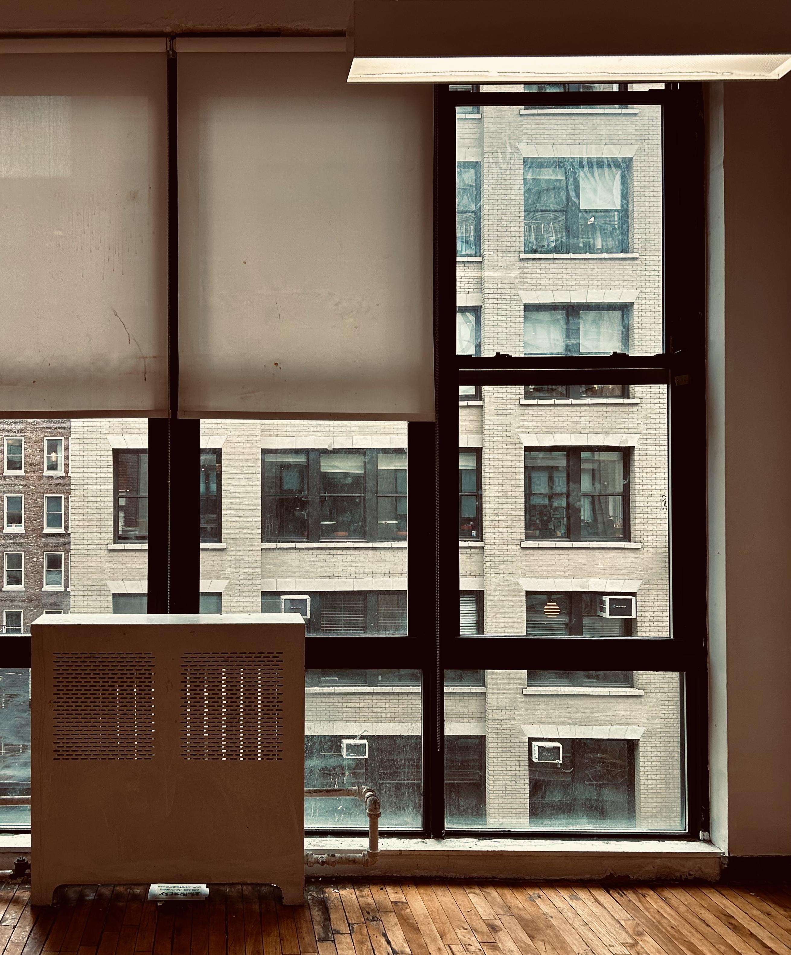 A color, medium-angle photograph in portrait orientation, an interior shot from a high-floor office building in Chelsea, Manhattan. The scene looks out through a large, industrial-style window toward a neighboring beige brick building. The window is divided into multiple panes by thick black metal frames, with two large white but aged roller shades partially drawn at the top, obscuring the upper left view. In the foreground, a vintage, cream-colored metal radiator cover sits on a classic light-wood parquet floor. A small section of exposed silver piping runs along the base of the window. Outside, the facade of the facing building dominates the view, featuring rows of large, uniform windows that reflect the overcast sky. Some of the windows across the way reveal glimpses of residential life, and several window-unit air conditioners are visible, tucked into the lower frames. To the far left, a sliver of a darker red-brick building provides a color contrast. The lighting is soft and muted, casting a neutral, slightly warm tone over the wooden floors and the weathered surfaces of the urban architecture.