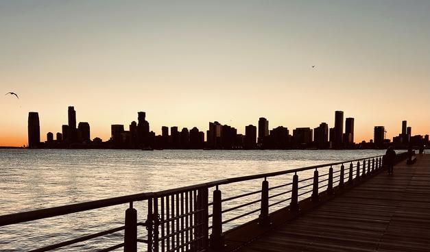 A color, wide-angle, silhouette-style photograph that captures the skyline of Jersey City from the wooden boards of Pier 45 in Manhattan during sunset. The foreground is dominated by a dark wooden boardwalk on the right, bordered by a metallic railing with vertical posts that recede into the distance. The calm waters of the Hudson River fill the midground, reflecting the soft, pale yellow and orange glow of the twilight sky. The horizon is a stark black silhouette of dense skyscrapers, with the prominent 99 Hudson Street and Goldman Sachs Tower standing out against the fading light. The sky transitions from a deep, warm orange near the horizon to a pale, hazy cream color towards the top. Two birds are visible in flight; one is soaring in the upper left corner with its wings spread wide, while another smaller bird appears as a tiny speck in the upper right. The overall mood is serene and cinematic, utilizing high contrast to emphasize the architectural outlines of the city against the natural gradient of the setting sun. Two people walk along the far right edge of the pier, adding a sense of scale and quiet solitude to the urban landscape.