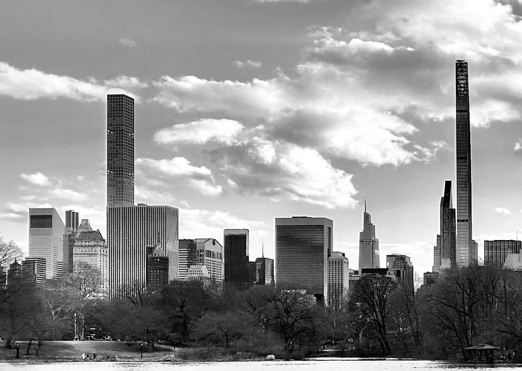A noir, wide-angle photograph in landscape orientation that captures the Manhattan skyline as seen from Central Park during the winter. In the foreground, a dark, calm body of water reflects a hint of the sky, bordered by a shoreline where several small figures of people are walking. The middle ground is filled with a dense thicket of bare, leafless deciduous trees, their intricate dark branches creating a stark contrast against the buildings. A small wooden gazebo sits nestled among the trees on the far right bank, adding a quiet, scenic element to the park landscape. The background is dominated by a row of towering skyscrapers under a bright sky filled with voluminous, textured clouds. Two exceptionally thin "pencil towers" bookend the composition: 432 Park Avenue on the left with its distinct grid-like facade, and 111 West 57th Street on the right, tapering sharply toward the top. Between them, a mix of modern glass towers and classic architectural styles, including the slanted roof of the Citigroup Center, creates a jagged and iconic horizon line. The lack of color emphasizes the dramatic lighting, casting deep shadows across the park and highlighting the reflective surfaces of the urban architecture.