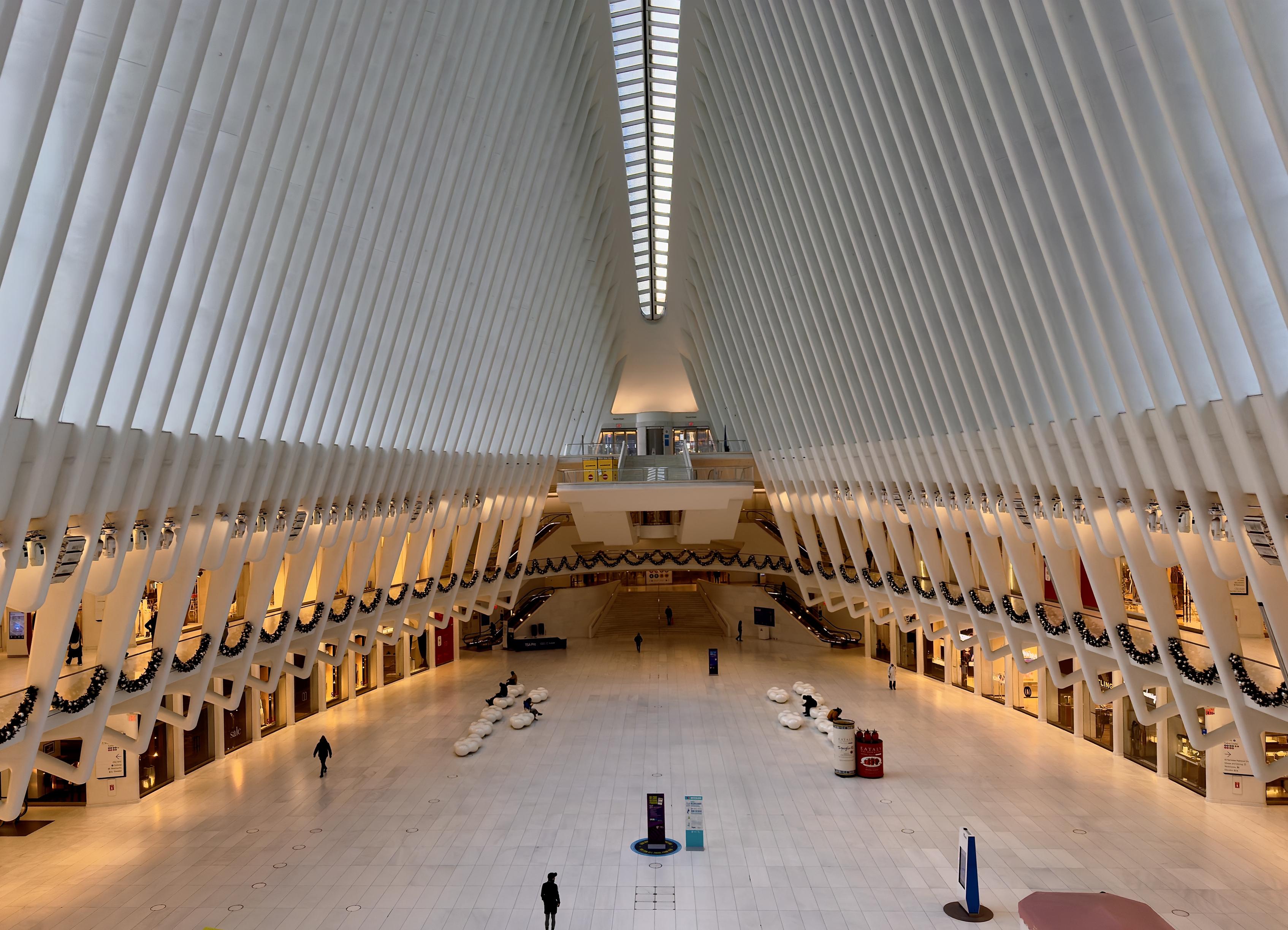 A color, wide-angle photograph taken from an elevated perspective looking down the main hall of the Oculus at the World Trade Center in New York City. The architectural space is characterized by a massive, white ribbed ceiling that curves upward to a central skylight, resembling the skeletal structure of a bird’s wings or a whale's ribcage. The symmetry of the white steel beams creates a striking sense of verticality and light. The polished white marble floor below reflects the bright overhead morning lighting, and a very few scattered pedestrians walk across the vast, open expanse. In the mid-ground, rows of white, circular seating pods are arranged for visitors. The transition between the lower retail levels and the soaring ceiling is marked by festive holiday decorations. Thick green evergreen garlands, draped in uniform scallops, are hung along the balconies of the second level, on both the left and right sides of the hall. The garlands are accented with small, silver ornaments and warm lights, providing a sharp organic contrast to the sharp, futuristic lines of the white architecture. In the far distance, a mezzanine level features additional holiday greenery and a small yellow kiosk, while the very back of the hall leads toward the subway entrances and further terminal corridors.
