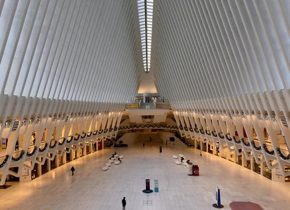 A color, wide-angle photograph taken from an elevated perspective looking down the main hall of the Oculus at the World Trade Center in New York City. The architectural space is characterized by a massive, white ribbed ceiling that curves upward to a central skylight, resembling the skeletal structure of a bird’s wings or a whale's ribcage. The symmetry of the white steel beams creates a striking sense of verticality and light. The polished white marble floor below reflects the bright overhead morning lighting, and a very few scattered pedestrians walk across the vast, open expanse. In the mid-ground, rows of white, circular seating pods are arranged for visitors. The transition between the lower retail levels and the soaring ceiling is marked by festive holiday decorations. Thick green evergreen garlands, draped in uniform scallops, are hung along the balconies of the second level, on both the left and right sides of the hall. The garlands are accented with small, silver ornaments and warm lights, providing a sharp organic contrast to the sharp, futuristic lines of the white architecture. In the far distance, a mezzanine level features additional holiday greenery and a small yellow kiosk, while the very back of the hall leads toward the subway entrances and further terminal corridors.