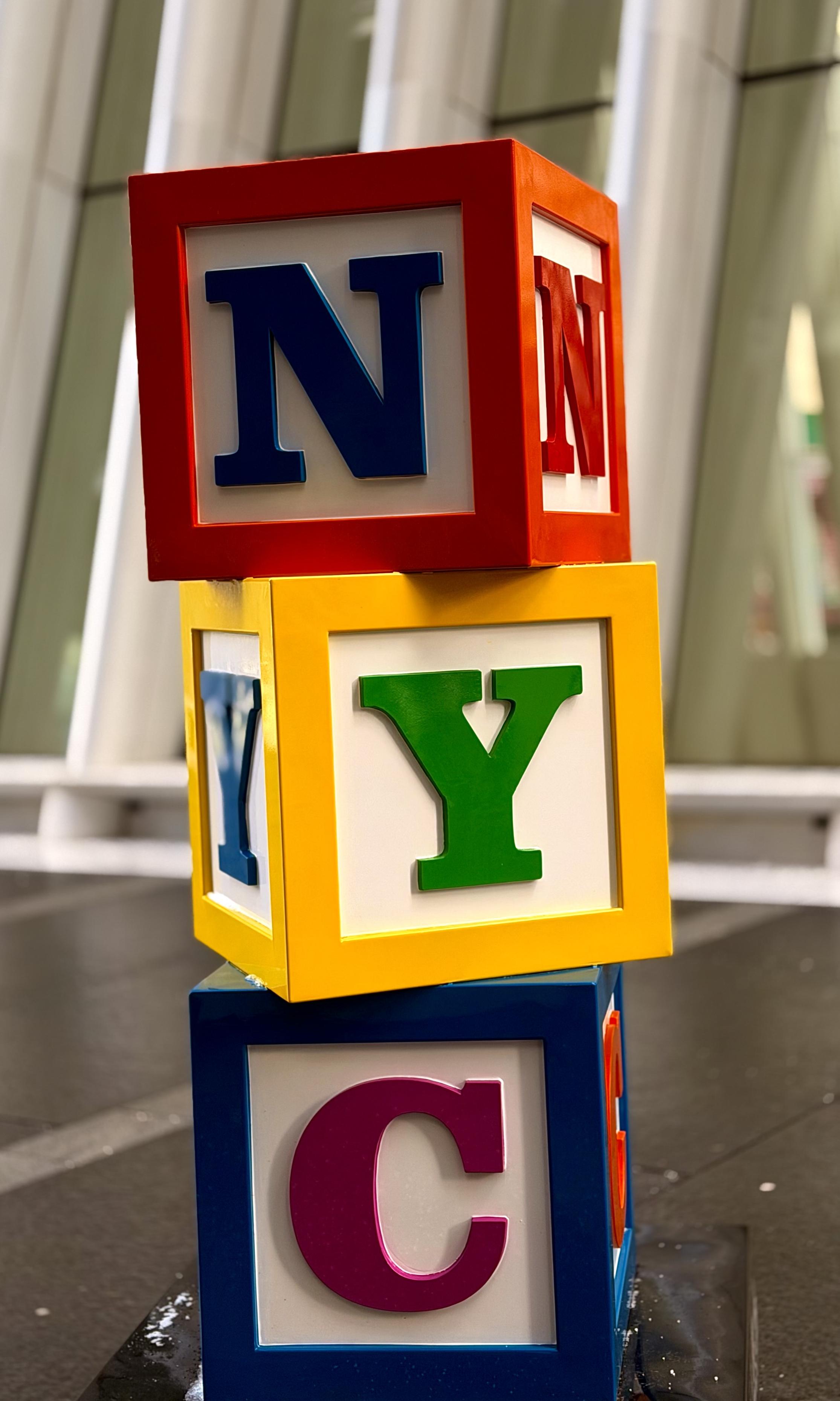 A color photograph in portrait orientation that captures a vibrant public art installation located at the South Oculus Plaza in New York City. The sculpture, part of the "Rewind" series by artist WhIsBe, consists of three oversized, stacked alphabet blocks that spell out "NYC" vertically. The top block is red with a blue letter "N," the middle is yellow with a green letter "Y," and the bottom is blue with a pinkish-purple letter "C." Each block features deeply recessed white panels that make the raised, colorful letters pop, mimicking the classic aesthetic of traditional wooden toy blocks but on a massive scale. The sculpture is positioned on a dark, reflective pedestal, which subtly mirrors the bright colors of the blocks in the early morning sunlight. In the background, the soft-focus, white vertical ribs of the Oculus transportation hub rise into the air, providing a clean, modern contrast to the playful, primary colors of the artwork.