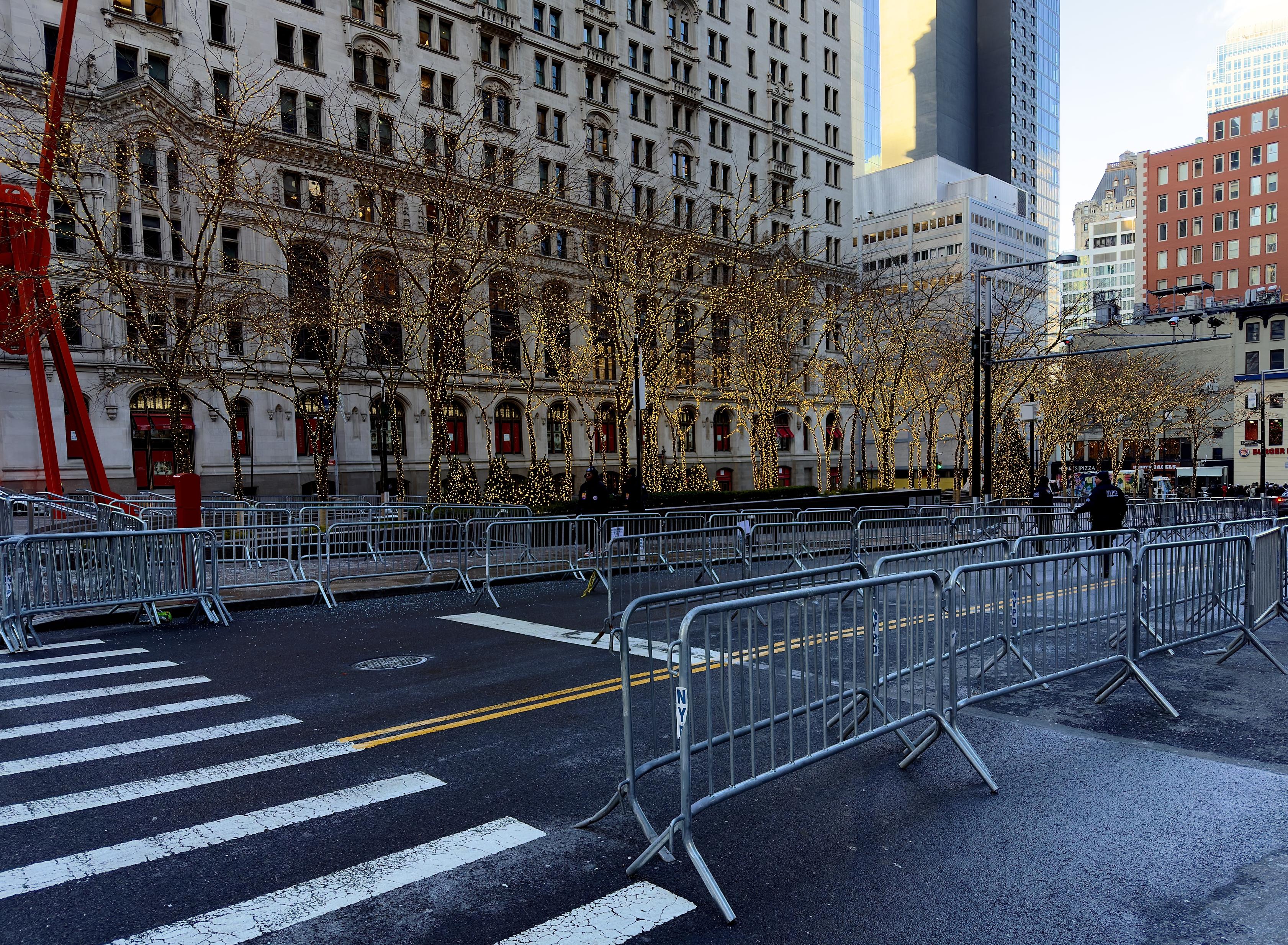 A color photograph that captures a cold morning in Lower Manhattan at Zuccotti Park, where security preparations are underway for the inauguration of New York City's 112th Mayor, Zohran Mamdani, on January 1, 2026. A wide crosswalk with thick white stripes leads into the frame from the bottom left. Several rows of silver metal barriers are positioned across the street, some with "NYPD" stenciled in blue on the vertical legs. Two NYPD officers in dark blue uniforms and high-visibility vests are visible in the distance on the right, managing the setup. To the left and center, the park is filled with rows of honey-locust trees, their trunks and branches wrapped in thousands of glowing warm-white fairy lights. Nestled among the trees is the bottom portion of the "Joie de Vivre" sculpture—a massive, bright red, tripod-like steel structure by artist Mark di Suvero. The ground appears damp, suggesting recent rain or a morning cleaning. The scene is quiet but industrious, capturing the "calm before the storm" as the city prepares for the public block party and swearing-in ceremony at nearby City Hall.