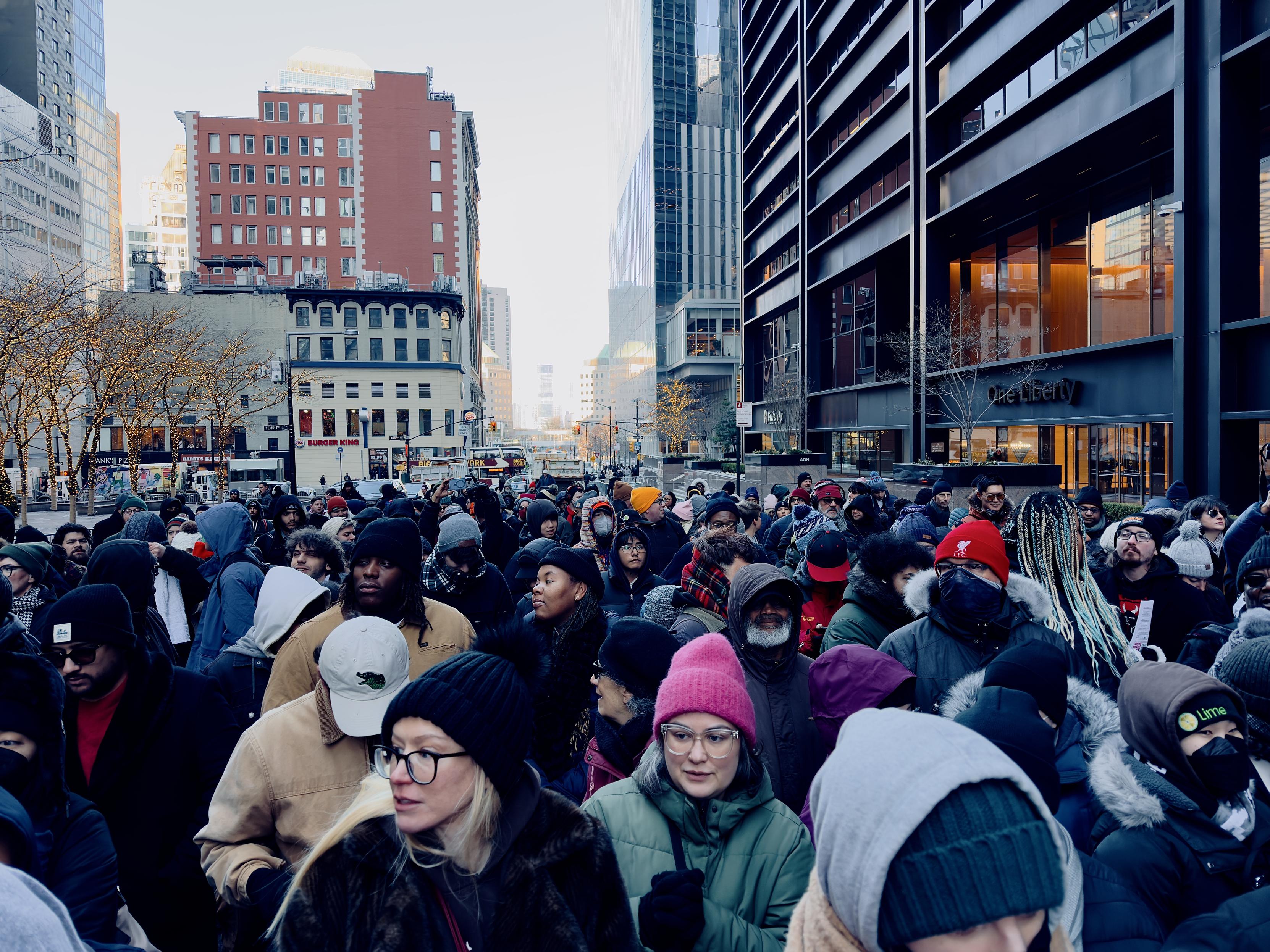 A color, ground-level, medium-angle photograph captures a dense, diverse crowd of people bundled in winter clothing, standing tightly packed on a city street waiting to enter the security screening for the mayoral inauguration in New York City today. In the foreground, individuals wear a variety of beanies, hooded parkas, and glasses, with their expressions ranging from patient to expectant. A woman in a bright pink beanie and another in a black knit hat and glasses are prominent in the lower-middle frame. The crowd fills the street, stretching back into the distance toward an intersection, creating a sea of dark coats punctuated by occasional splashes of color from hats and bags. The perspective looks down Liberty Street toward Church Street in a bustling urban environment. To the right, the sleek, dark architectural facade of the "One Liberty" building rises sharply, its glass windows reflecting the soft, cool daylight. On the left, older stone buildings house storefronts like a Burger King, while bare-branched trees decorated with small white fairy lights line the sidewalk. The clear, pale sky hangs above the narrow canyon created by the tall buildings, and a few city buses and traffic signals are visible in the far background where the crowd eventually thins.