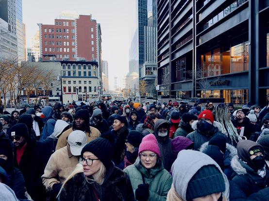 A color, ground-level, medium-angle photograph captures a dense, diverse crowd of people bundled in winter clothing, standing tightly packed on a city street waiting to enter the security screening for the mayoral inauguration in New York City today. In the foreground, individuals wear a variety of beanies, hooded parkas, and glasses, with their expressions ranging from patient to expectant. A woman in a bright pink beanie and another in a black knit hat and glasses are prominent in the lower-middle frame. The crowd fills the street, stretching back into the distance toward an intersection, creating a sea of dark coats punctuated by occasional splashes of color from hats and bags. The perspective looks down Liberty Street toward Church Street in a bustling urban environment. To the right, the sleek, dark architectural facade of the "One Liberty" building rises sharply, its glass windows reflecting the soft, cool daylight. On the left, older stone buildings house storefronts like a Burger King, while bare-branched trees decorated with small white fairy lights line the sidewalk. The clear, pale sky hangs above the narrow canyon created by the tall buildings, and a few city buses and traffic signals are visible in the far background where the crowd eventually thins.