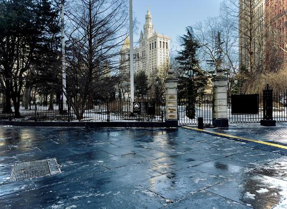 A color, wide-angle photograph of the urban landscape along Broadway in front of City Hall Park in Manhattan. It is a bitter cold, and the light blue morning sky is illuminating the scene. In the foreground of the image, the dark, slate-grey stone pavers of the sidewalk appear slick and reflective, dotted with patches of melting snow and ice. A metal drainage grate is embedded in the ground to the left, and a bright yellow tactile paving strip runs horizontally across the path toward the right. The damp surface captures the pale blue light of the winter sky. Beyond a black wrought-iron fence, the skeletal branches of winter-bare trees fill the mid-ground, partially obscuring the historic buildings in the distance. A sign with white text reads “City Hall Park”. Though a Jumbotron screen is peeking through in the distance from the public inauguration of Mayor Zohran Mamdani taking place just a block away on the steps of City Hall, this corner is still. 