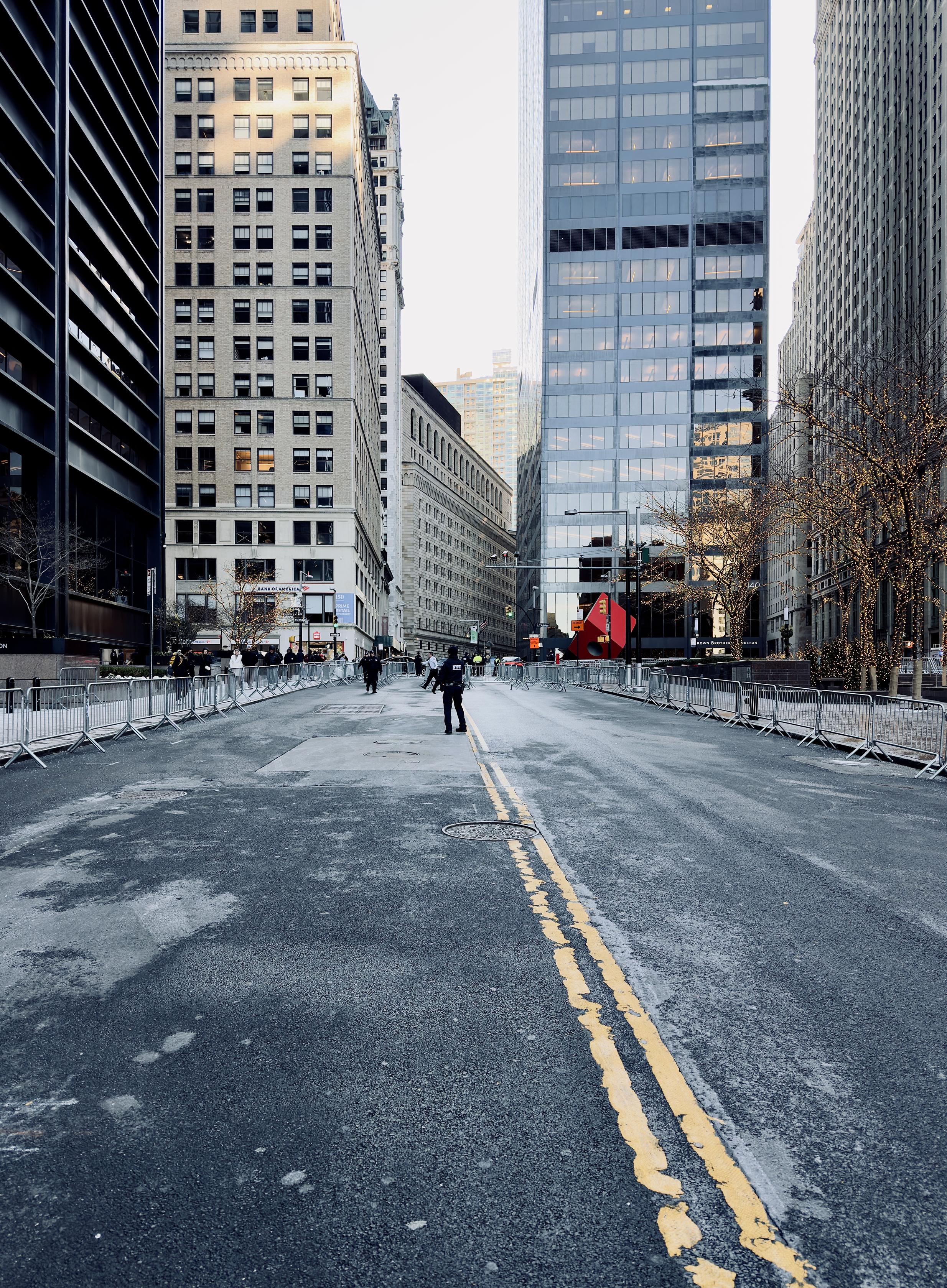 A color photograph in landscape orientation captures a wide, multi-lane street in Lower Manhattan as seen from a low-angle perspective, completely cleared of vehicle traffic and lined with metal police barricades. The asphalt is weathered with light gray patches and a double yellow line that curves slightly toward the right. In the distance, a solitary police officer stands near the center of the road facing away from the camera. Many other police officers are in small groups dispersed along the intersection with Broadway. The street is flanked by tall, modern glass skyscrapers and historic stone buildings. The barricades form a continuous silver border on both sides of the roadway. To the right, several leafless trees are wrapped in warm, glowing fairy lights. In the background, a large, iconic red cube sculpture sits at the intersection, serving as a bright focal point against the gray tones of the street. The sky above is a clear, pale blue, casting a soft, desaturated even light over the scene.
