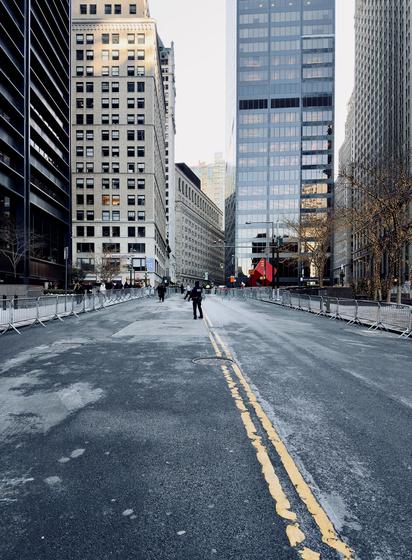 A color photograph in landscape orientation captures a wide, multi-lane street in Lower Manhattan as seen from a low-angle perspective, completely cleared of vehicle traffic and lined with metal police barricades. The asphalt is weathered with light gray patches and a double yellow line that curves slightly toward the right. In the distance, a solitary police officer stands near the center of the road facing away from the camera. Many other police officers are in small groups dispersed along the intersection with Broadway. The street is flanked by tall, modern glass skyscrapers and historic stone buildings. The barricades form a continuous silver border on both sides of the roadway. To the right, several leafless trees are wrapped in warm, glowing fairy lights. In the background, a large, iconic red cube sculpture sits at the intersection, serving as a bright focal point against the gray tones of the street. The sky above is a clear, pale blue, casting a soft, desaturated even light over the scene.