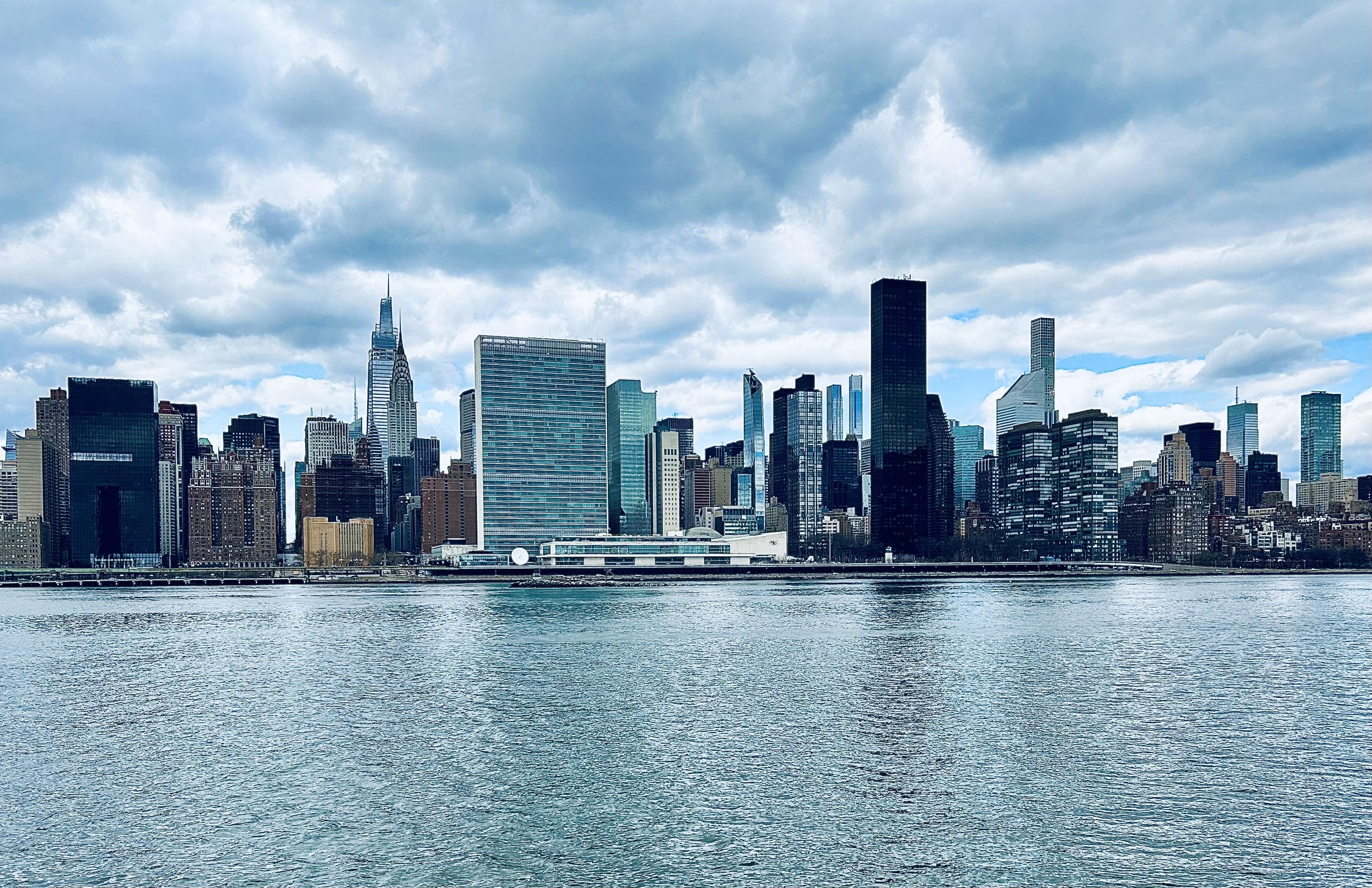 A color, wide-angle photograph in landscape orientation that captures the Midtown Manhattan skyline as viewed from across the East River in Long Island City, Queens on a partly cloudy winter afternoon. The foreground is dominated by the textured, shimmering blue-grey water of the river, which transitions into the dark shoreline of Manhattan. The skyline features a dense array of skyscrapers under a dramatic, overcast sky filled with heavy, layered grey and white clouds. Notable landmarks are visible, including the slender, tiered spire of the Chrysler Building and the modern, pointed peak of One Vanderbilt. Centrally located is the distinctive, rectangular glass slab of the United Nations Secretariat Building, reflecting the cool tones of the sky. The composition highlights a mix of architectural styles, ranging from dark, monolithic rectangular towers to slim, ultra-modern residential "pencil towers" like 432 Park Avenue in the distant background. To the right of the United Nations, several glassy residential buildings line the waterfront, while the far left shows the dark, blocky silhouette of the Solow Building. The color palette is cool and somewhat desaturated, characterized by steely blues, deep greys, and the various shades of glass and steel from the buildings. The overall atmosphere is serene yet powerful, emphasizing the scale and density of the urban landscape against the expansive sky.