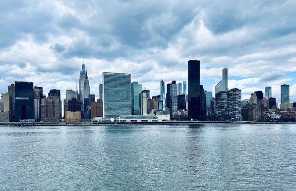 A color, wide-angle photograph in landscape orientation that captures the Midtown Manhattan skyline as viewed from across the East River in Long Island City, Queens on a partly cloudy winter afternoon. The foreground is dominated by the textured, shimmering blue-grey water of the river, which transitions into the dark shoreline of Manhattan. The skyline features a dense array of skyscrapers under a dramatic, overcast sky filled with heavy, layered grey and white clouds. Notable landmarks are visible, including the slender, tiered spire of the Chrysler Building and the modern, pointed peak of One Vanderbilt. Centrally located is the distinctive, rectangular glass slab of the United Nations Secretariat Building, reflecting the cool tones of the sky. The composition highlights a mix of architectural styles, ranging from dark, monolithic rectangular towers to slim, ultra-modern residential "pencil towers" like 432 Park Avenue in the distant background. To the right of the United Nations, several glassy residential buildings line the waterfront, while the far left shows the dark, blocky silhouette of the Solow Building. The color palette is cool and somewhat desaturated, characterized by steely blues, deep greys, and the various shades of glass and steel from the buildings. The overall atmosphere is serene yet powerful, emphasizing the scale and density of the urban landscape against the expansive sky.