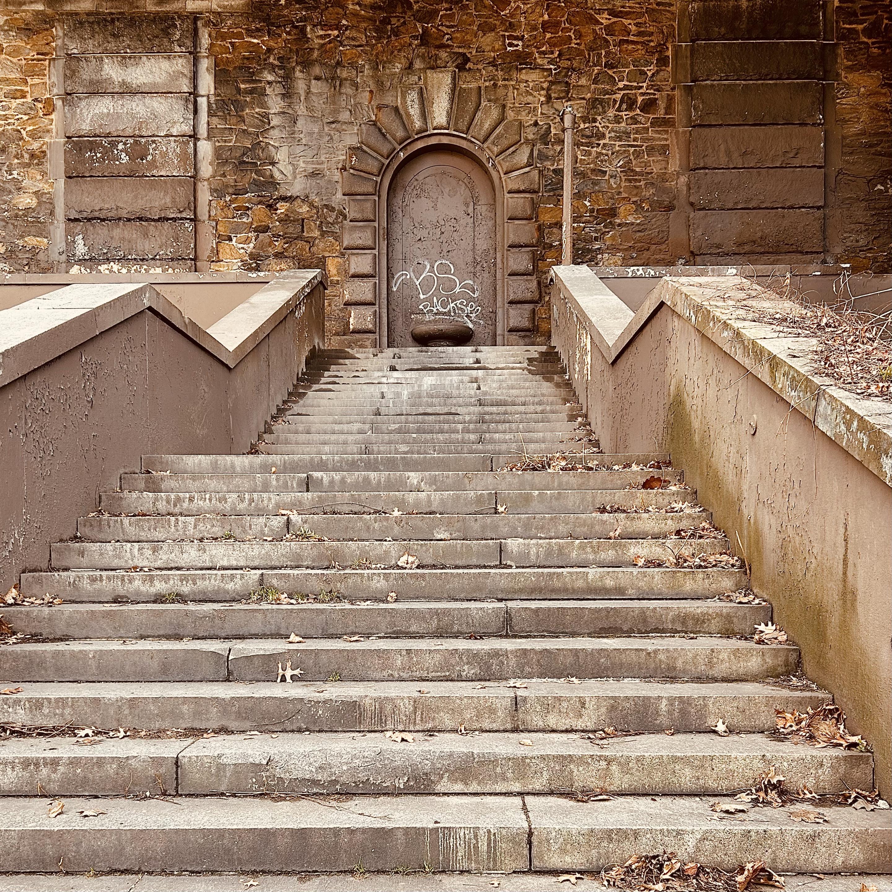 A color, medium-angle photograph in square aspect ratio. An eye-level shot that captures a weathered stone staircase leading up to a grand, arched doorway set into a rustic stone wall. The steps are wide and made of grey, rectangular stone slabs, scattered with dried brown leaves and debris, indicating the winter setting. Flanking the stairs are thick, angled concrete banisters in a muted brownish-mauve tone, which show signs of peeling paint and wear. The overall color palette is earthy and desaturated, dominated by shades of tan, brown, and grey. At the top of the stairs, a heavy metal door is recessed into a deep archway framed by prominent, rusticated stone blocks. The door is covered in white graffiti tags, most notably the letters "BS" and "BACKSEE." To either side of the central arch, the wall features large, rectangular decorative panels made of horizontal stone courses, creating a sense of classical architectural order despite the structure's weathered and neglected state. A thin metal pipe runs vertically along the wall to the right of the door, and a few dry, leafless branches peek over the top of the right-hand banister.