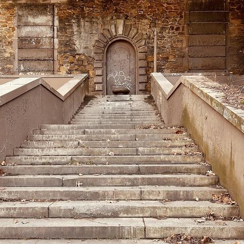 A color, medium-angle photograph in square aspect ratio. An eye-level shot that captures a weathered stone staircase leading up to a grand, arched doorway set into a rustic stone wall. The steps are wide and made of grey, rectangular stone slabs, scattered with dried brown leaves and debris, indicating the winter setting. Flanking the stairs are thick, angled concrete banisters in a muted brownish-mauve tone, which show signs of peeling paint and wear. The overall color palette is earthy and desaturated, dominated by shades of tan, brown, and grey. At the top of the stairs, a heavy metal door is recessed into a deep archway framed by prominent, rusticated stone blocks. The door is covered in white graffiti tags, most notably the letters "BS" and "BACKSEE." To either side of the central arch, the wall features large, rectangular decorative panels made of horizontal stone courses, creating a sense of classical architectural order despite the structure's weathered and neglected state. A thin metal pipe runs vertically along the wall to the right of the door, and a few dry, leafless branches peek over the top of the right-hand banister.