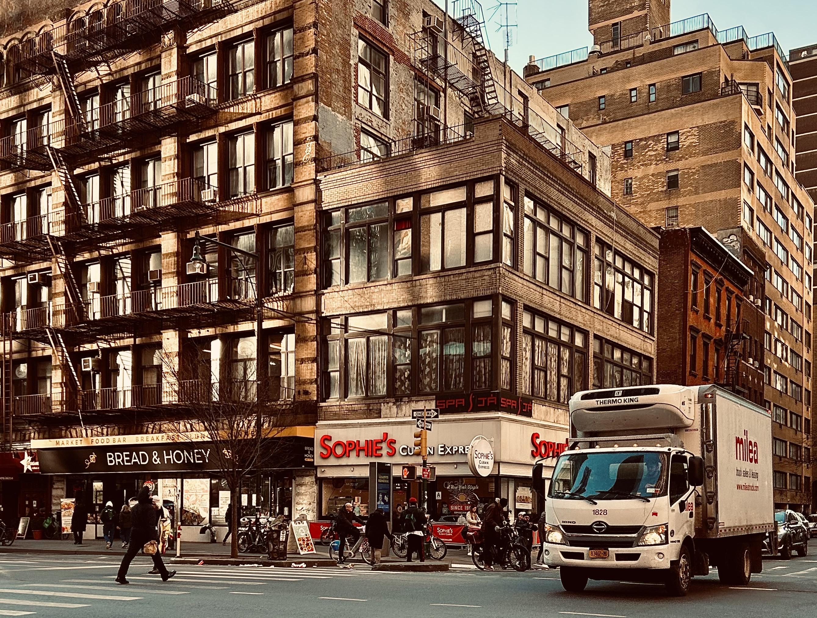 A color, medium-angle photograph of the street scene in Midtown Manhattan near dusk on a clear winter day. The architecture is dominated by multi-story, weathered brick buildings featuring industrial-style windows and intricate black metal fire escapes that zigzag down the facades. On the ground floor, prominent storefronts include "Bread & Honey," a market and food bar with a sleek black awning, and "Sophie’s Cuban Cuisine," marked by vibrant red lettering and a circular white sign. Long, dramatic shadows stretch across the asphalt, cast by the low-angled sun, while the warm, sepia-toned light reflects off the glass panes and masonry, creating a high-contrast atmosphere. In the foreground, a large white Milea box truck with a Thermo King refrigeration unit and Hino branding drives through the intersection from right to left. Pedestrians bundled in heavy winter coats, hats, and scarves navigate the crosswalks and sidewalks, including a person in a black parka walking briskly toward the left. Several delivery cyclists and bicycles are clustered near the corner of the Sophie’s storefront, leaning against traffic poles. In the background, taller residential and office towers rise into a clear sky, their tan and brown brickwork catching the fading daylight.