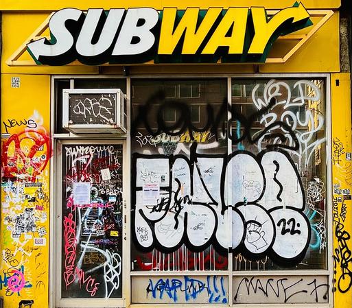 A color photograph in square aspect ratio. The focal point is a close-up, front-facing shot showing a closed Subway sandwich shop with its storefront almost entirely covered in graffiti and street art. A large, yellow and white Subway logo is mounted on a yellow facade above a glass window and door. The glass is heavily layered with various styles of graffiti, including large, bubbly white "throw-up" letters outlined in black, spindly black tags, and colorful scribbles in pink, red, and blue. Stickers and small flyers are plastered across the yellow door frame and the glass. An old, rusted window air conditioning unit is installed above the glass door on the left, also marked with white spray paint. Through the relatively clear sections of the window, the faint reflection of brick buildings across the street are visible. At the bottom of the window, more linear black and blue tags stretch across the glass pane. The overall scene captures a gritty, abandoned look, contrasting the bright, corporate branding of the restaurant with the chaotic, dense layering of local street culture.