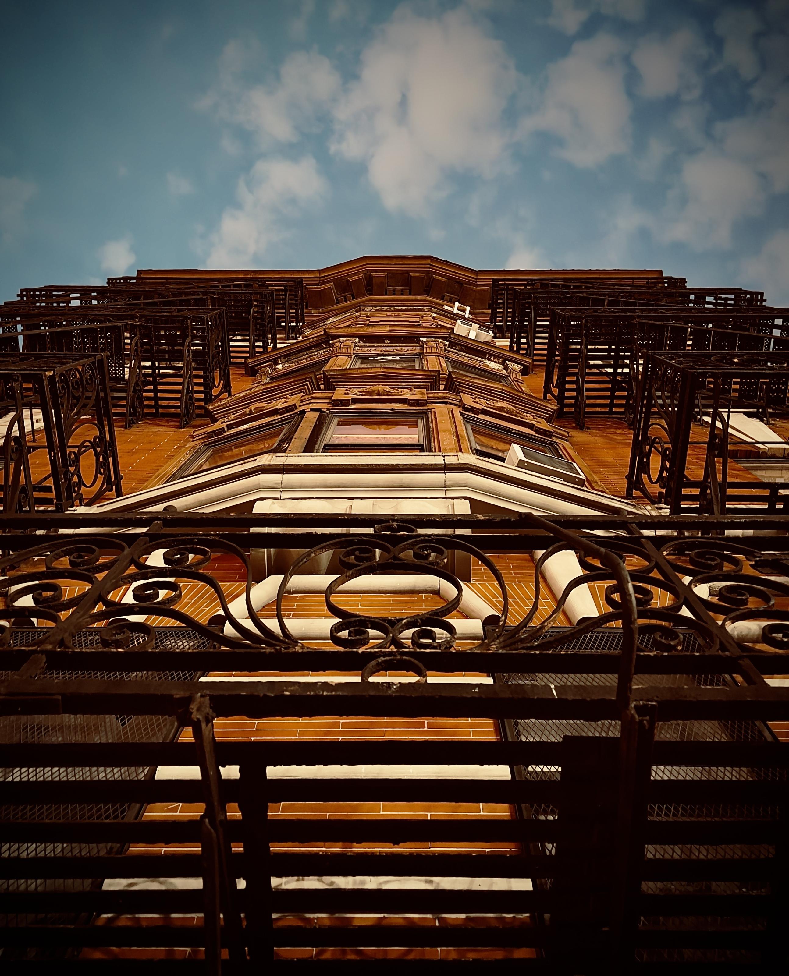 A color, low-angle photograph in portrait orientation. A dramatic shot looking straight up the facade of a classic Upper West Side brownstone toward an early evening twilight sky. The building is constructed of warm, orange-toned brick with elaborate architectural details, including protruding bay windows and white stone lintels. Intricate, black wrought-iron fire escapes and balconies dominate the foreground and sides of the frame, their swirling decorative patterns creating a complex silhouette against the building’s face. The perspective creates a strong sense of verticality, narrowing as the building rises toward the top of the frame. The sky above is a deep, moody blue, filled with scattered, soft white clouds that are illuminated by the fading evening light. A slight vignette around the edges of the image focuses attention on the central architectural details, emphasizing the contrast between the dark ironwork, the glowing brick, and the expansive sky.