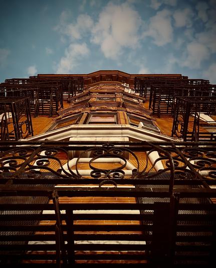 A color, low-angle photograph in portrait orientation. A dramatic shot looking straight up the facade of a classic Upper West Side brownstone toward an early evening twilight sky. The building is constructed of warm, orange-toned brick with elaborate architectural details, including protruding bay windows and white stone lintels. Intricate, black wrought-iron fire escapes and balconies dominate the foreground and sides of the frame, their swirling decorative patterns creating a complex silhouette against the building’s face. The perspective creates a strong sense of verticality, narrowing as the building rises toward the top of the frame. The sky above is a deep, moody blue, filled with scattered, soft white clouds that are illuminated by the fading evening light. A slight vignette around the edges of the image focuses attention on the central architectural details, emphasizing the contrast between the dark ironwork, the glowing brick, and the expansive sky.