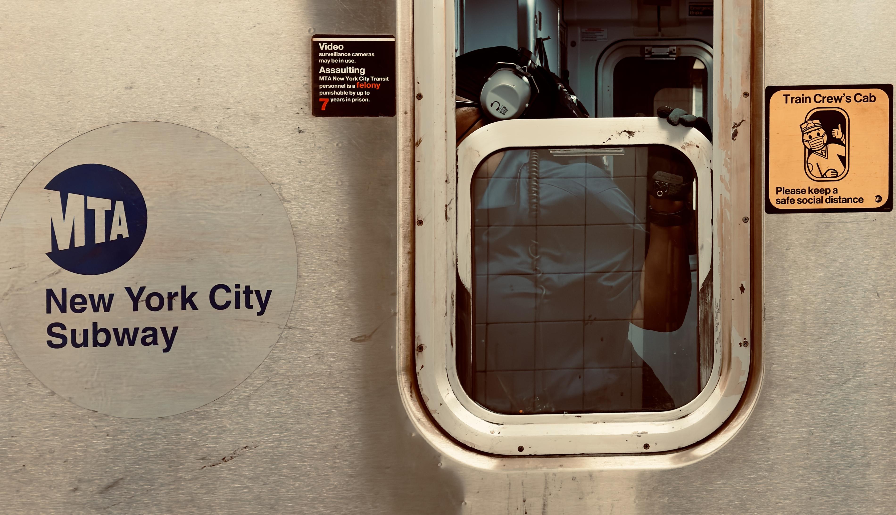 A color photograph in landscape orientation. A close-up, eye-level shot captures the exterior of a stainless steel New York City Subway car. On the left, a large, faded circular decal features the blue "MTA" logo above the words "New York City Subway" in a dark sans-serif font. To the right, a rectangular window with rounded corners reveals a glimpse into the train crew's cab. Inside, a person wearing a blue shirt and large white over-ear hearing protection is visible, their back partially turned to the window. A black gloved hand rests on the upper frame of the window, and a coiled black handset cord hangs against the interior glass. The train’s metallic surface is marked by various signs and textures. Above the cab window, a small black sign warns that assaulting transit personnel is a felony punishable by up to seven years in prison. To the right of the window, a yellow square sign labeled "Train Crew's Cab" features an illustration of a masked crew member giving a thumbs-up, accompanied by a request to maintain a safe social distance. The overall lighting is warm and slightly desaturated, emphasizing the industrial, weathered texture of the subway car’s brushed metal exterior and the various rivets holding the window frame in place.