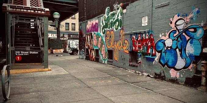 A color, wide-angle photograph that captures a gritty urban sidewalk scene in the Inwood neighborhood of Manhattan, right outside the 215th Street subway station entrance. On the left, a traditional dark green metal subway entrance with ornate wrought-iron details houses a staircase leading down; a black sign with a red "1" circle identifies it as the station for the 1 train toward South Ferry. The background shows the neighborhood’s character with a white van covered in graffiti parked near a storefront and the elevated train tracks casting shadows over the street. The ground is made of large gray concrete sidewalk slabs, marked with faint orange utility graffiti. Dominating the right side of the frame is a long brick wall completely covered in layers of vibrant, multi-colored street art and bubble-letter graffiti. Bold tags in shades of blue, red, green, and yellow pop against the gray brick, with names like "Clyde" and "Bama" clearly visible. The lighting is overcast and slightly moody, emphasizing the high contrast between the colorful mural and the dark, industrial architecture of the subway station.