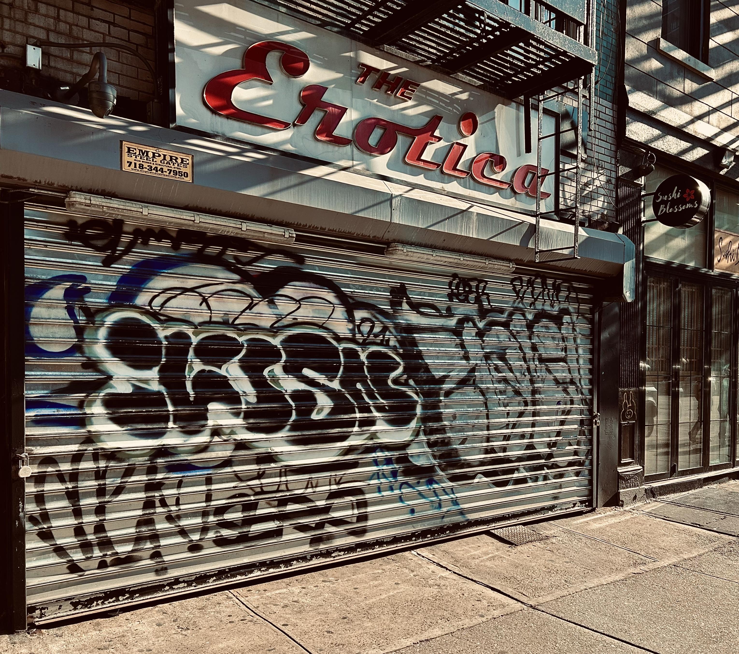 A color photograph in square aspect ratio. An eye-level, medium-angle capture of the street scene along Eighth Avenue in the Chelsea neighborhood of Manhattan. The focal point of the image is a weathered storefront. Fromvthe sidewalk facing a closed, silver metal security gate heavily covered in black and blue graffiti tags and "throw-ups." Above the gate, a large vintage sign reads "THE Erotica" in stylized, red cursive lettering against a white background. Shadows from a fire escape overhead cast diagonal, dark stripes across the sign and the building's facade. To the left, a small yellow sticker for "Empire Steel Gates" is visible, and a security camera is mounted on the brickwork. To the right of the storefront, a modern restaurant called "Sushi Blossoms" provides a sharp contrast. It features a clean, glass-paned facade and a small, circular black sign with a red flower logo. The sidewalk in the foreground is made of light gray concrete slabs, partially shaded by the surrounding buildings. The overall color palette is somewhat desaturated with high contrast, leaning into an urban, gritty aesthetic.