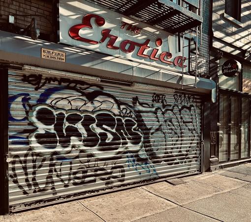 A color photograph in square aspect ratio. An eye-level, medium-angle capture of the street scene along Eighth Avenue in the Chelsea neighborhood of Manhattan. The focal point of the image is a weathered storefront. Fromvthe sidewalk facing a closed, silver metal security gate heavily covered in black and blue graffiti tags and "throw-ups." Above the gate, a large vintage sign reads "THE Erotica" in stylized, red cursive lettering against a white background. Shadows from a fire escape overhead cast diagonal, dark stripes across the sign and the building's facade. To the left, a small yellow sticker for "Empire Steel Gates" is visible, and a security camera is mounted on the brickwork. To the right of the storefront, a modern restaurant called "Sushi Blossoms" provides a sharp contrast. It features a clean, glass-paned facade and a small, circular black sign with a red flower logo. The sidewalk in the foreground is made of light gray concrete slabs, partially shaded by the surrounding buildings. The overall color palette is somewhat desaturated with high contrast, leaning into an urban, gritty aesthetic.