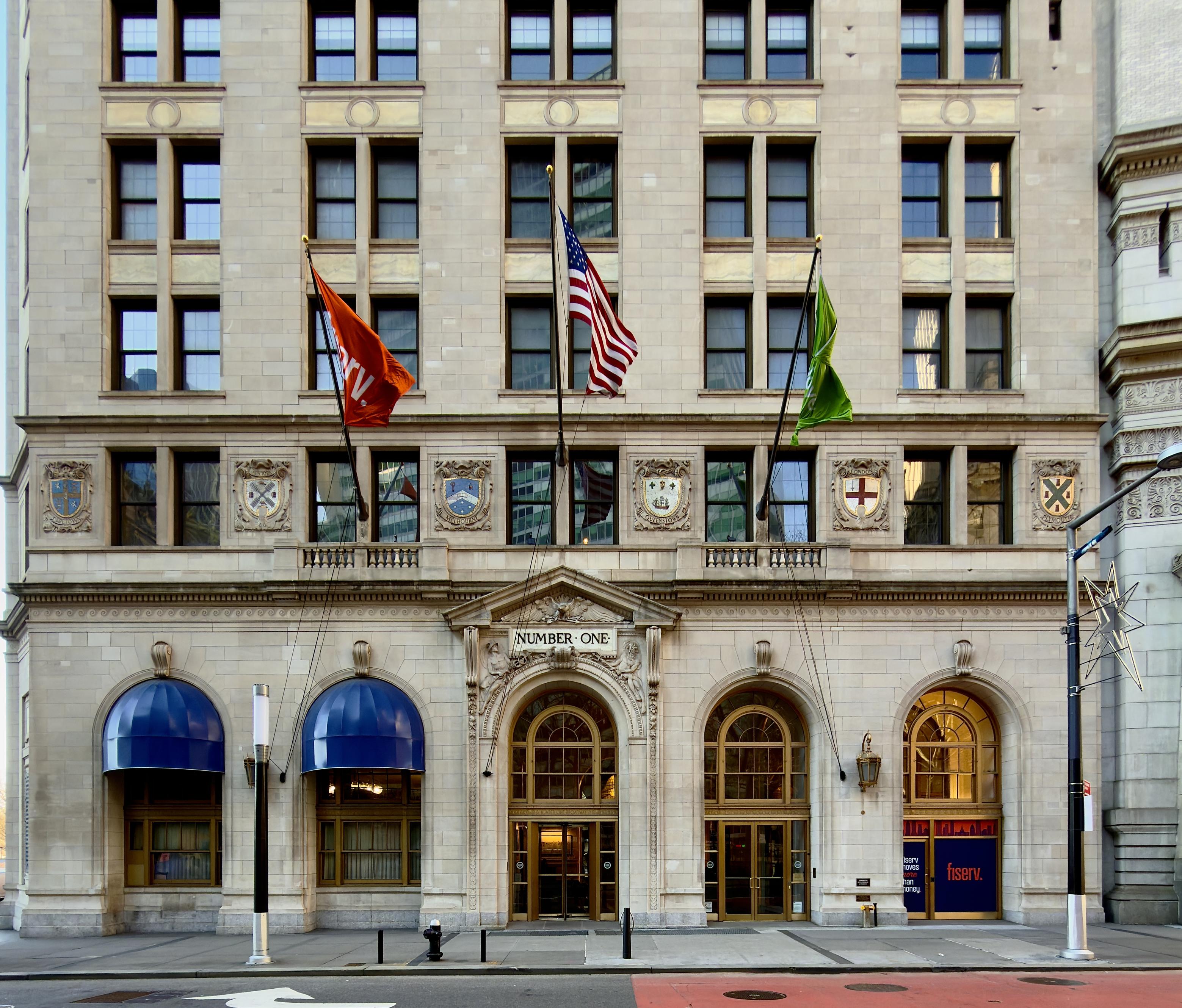 A color photograph in square aspect ratio that captures the facade of the historic International Mercantile Marine Building, built in 1884, located at 1 Broadway in the Financial District of Manhattan. The architecture is a grand example of Neoclassical design, featuring a light-colored stone exterior with deep-set windows and a series of arched entryways at the street level. Above the main central entrance, the words "NUMBER ONE" are carved into the stone. To the right, a large window displays the branding for Fiserv, featuring their signature orange logo against a blue background. The building is further decorated with several ornate stone crests and shields positioned between the first and second floors that honor port cities around the world, adding to its stately, historic character. The mid-section of the building features three large flagpoles extending over the sidewalk. The central pole flies the United States flag, flanked by an orange flag on the left and a lime green flag on the right. Below these flags, the street-level architecture includes two prominent bright blue rounded awnings over the windows to the left of the main entrance. The sidewalk in the foreground is clean and modern, equipped with black bollards, a fire hydrant, and a sleek street lamp decorated with a geometric star-shaped ornament. The overall perspective is a straight-on, symmetrical view that emphasizes the building’s monumental scale and architectural detail.