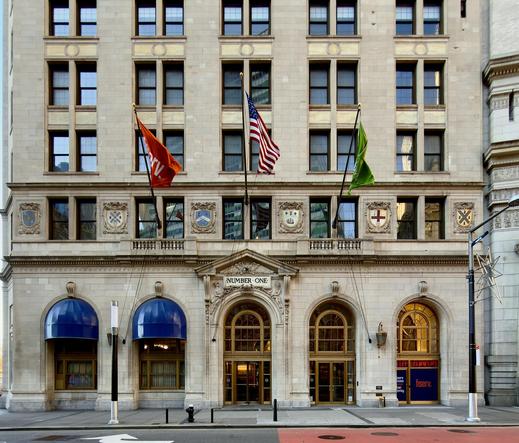 A color photograph in square aspect ratio that captures the facade of the historic International Mercantile Marine Building, built in 1884, located at 1 Broadway in the Financial District of Manhattan. The architecture is a grand example of Neoclassical design, featuring a light-colored stone exterior with deep-set windows and a series of arched entryways at the street level. Above the main central entrance, the words "NUMBER ONE" are carved into the stone. To the right, a large window displays the branding for Fiserv, featuring their signature orange logo against a blue background. The building is further decorated with several ornate stone crests and shields positioned between the first and second floors that honor port cities around the world, adding to its stately, historic character. The mid-section of the building features three large flagpoles extending over the sidewalk. The central pole flies the United States flag, flanked by an orange flag on the left and a lime green flag on the right. Below these flags, the street-level architecture includes two prominent bright blue rounded awnings over the windows to the left of the main entrance. The sidewalk in the foreground is clean and modern, equipped with black bollards, a fire hydrant, and a sleek street lamp decorated with a geometric star-shaped ornament. The overall perspective is a straight-on, symmetrical view that emphasizes the building’s monumental scale and architectural detail.