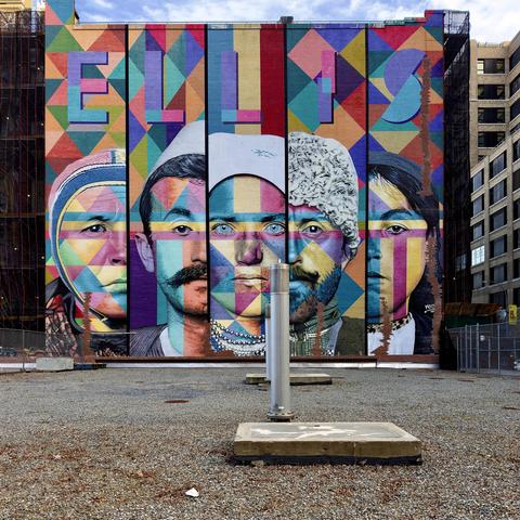 A color photograph in square aspect ratio that captures a vibrant, large-scale mural titled "ELLIS" which covers the side of a multi-story brick building, viewed from a gravel-covered lot in Manhattan's West Village. The mural features five stylized, realistic portraits of immigrants, inspired by historical Ellis Island archives. The faces are rendered with a mix of natural skin tones and a colorful, geometric diamond pattern that overlays the entire composition. Above the figures, the word "ELLIS" is written in large, bold, blue block letters. The background of the mural consists of a kaleidoscopic Harlequin pattern in bright shades of red, yellow, blue, and purple. In the foreground, a flat, grey gravel roof occupies the lower third of the frame, featuring a concrete base and a tall, cylindrical silver vent pipe standing vertically in the center. To the left of the mural, scaffolding and black netting shroud an adjacent building under construction, while to the right, a modern office building with large windows is visible. The mural itself is divided into five vertical segments by dark lines, giving it a paneled appearance. The lighting is bright and even, suggesting a clear day, highlighting the sharp contrast between the gritty industrial rooftop and the brilliant, multifaceted colors of the public art.
