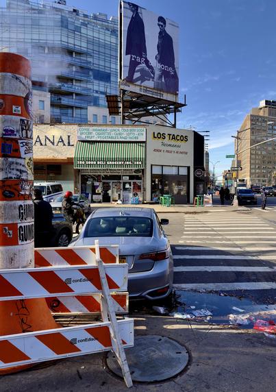 A color photograph in portrait orientation of the street scene in the Tribeca neighborhood of Manhattan. The scene is dominated by a mix of gritty urban textures and modern commercial elements under a clear blue sky. On the left foreground, a tall orange-and-white striped steam pipe and ConEdison construction barriers. Behind these, a silver sedan is parked near a crosswalk. The street level features a row of small businesses, most notably a storefront with a white sign where the first letter is obscured by the steam pipe, humorously making the word "CANAL" appear as "ANAL." Next to it is the green-and-white striped awning of the Soho Deli Grocery and a "Los Tacos Hermanos" restaurant. Above the shops, a massive "rag & bone NEW YORK" billboard towers over the intersection, while a modern glass skyscraper and a classic brick industrial building rise in the background. The sidewalk is scattered with light litter and a small puddle reflects the bright sunlight, while a few pedestrians and a person on a scooter move through the frame. The steam pipe is heavily weathered and covered in various street art stickers. The lighting is sharp and directional, casting long shadows across the asphalt and highlighting the contrast between the older, low-rise storefronts and the sleek, reflective glass of the newer architecture nearby.