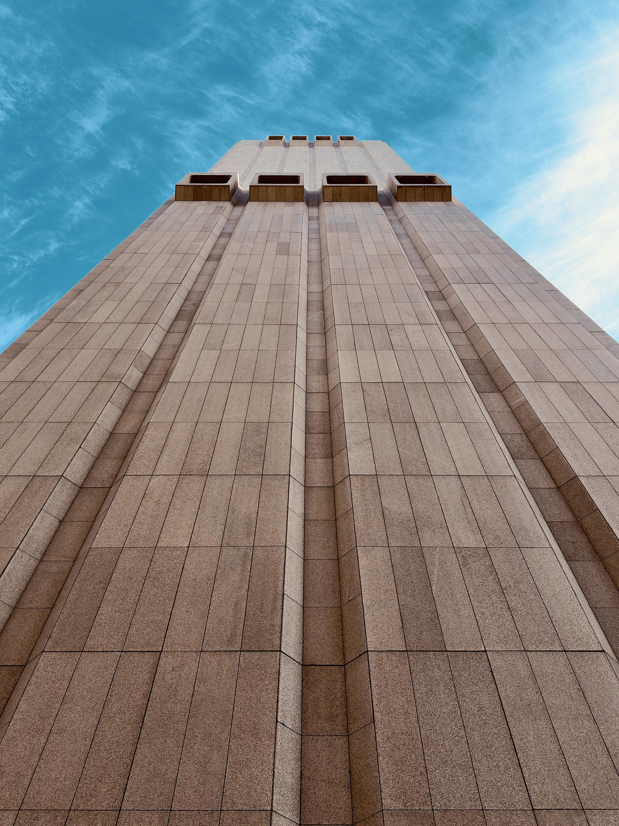 A color, low-angle photograph in portrait orientation that captures the windowless, Brutalist facade of the Long Lines Building at 33 Thomas Street in the Tribeca neighborhood of Manhattan. The building is rising dramatically against a bright blue sky with wispy white clouds. The structure is clad in textured, flame-cut Swedish granite panels in a warm hue, organized into deep vertical recessed channels that draw the eye upward. Near the top of the frame, large, rectangular concrete ventilation openings protrude from the flat surface like blocky, hollow balconies. The perspective creates a sense of immense scale and monolithic power, emphasizing the building's fortress-like, utilitarian design.