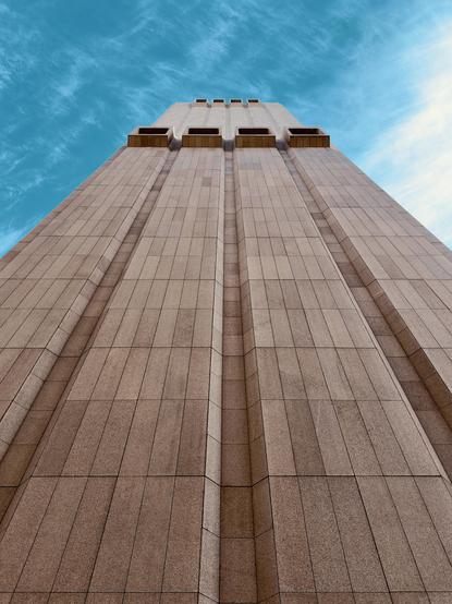 A color, low-angle photograph in portrait orientation that captures the windowless, Brutalist facade of the Long Lines Building at 33 Thomas Street in the Tribeca neighborhood of Manhattan. The building is rising dramatically against a bright blue sky with wispy white clouds. The structure is clad in textured, flame-cut Swedish granite panels in a warm hue, organized into deep vertical recessed channels that draw the eye upward. Near the top of the frame, large, rectangular concrete ventilation openings protrude from the flat surface like blocky, hollow balconies. The perspective creates a sense of immense scale and monolithic power, emphasizing the building's fortress-like, utilitarian design.