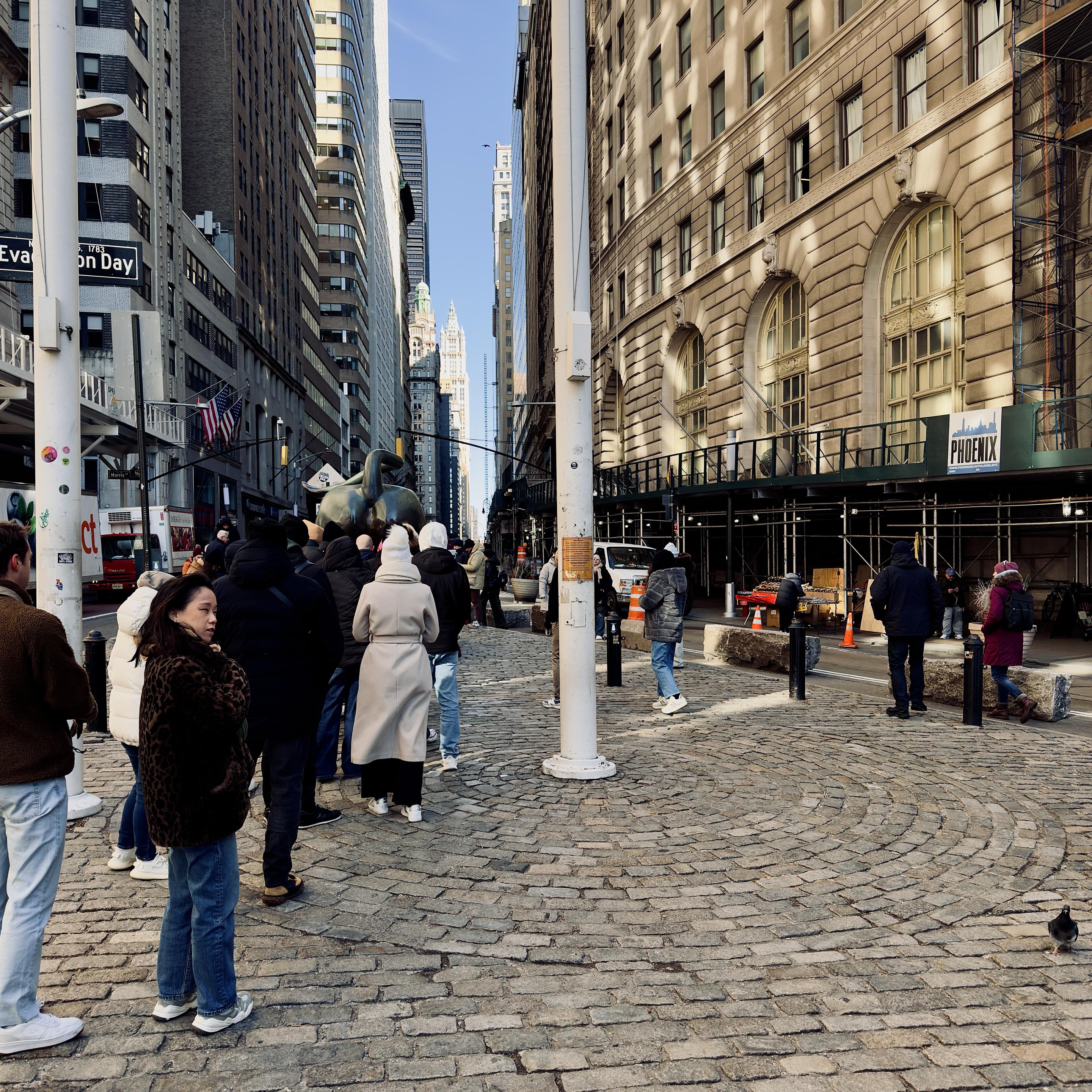 A color photograph in square aspect ratio of the street scene along Broadway in the Financial District of Manhattan. The image captures a crowd of people waiting in a long, organized line to see the Charging Bull sculpture. The perspective is from behind the crowd, looking north up Broadway. A diverse group of tourists, dressed in heavy winter coats, hats, and scarves, stand on the historic, fan-patterned cobblestone plaza. In the center-left, the bronze hindquarters of the bull are visible as people wait for their turn to take photos. To the right, a tall white utility pole stands in the foreground, while a small pigeon walks across the stones in the bottom right corner. The scene is framed by the towering, classic architecture of Lower Manhattan. On the right, a large stone building features grand arched windows and is partially covered by green construction scaffolding with a "PHOENIX" sign. On the left, a street sign marks "Evacuation Day" and "Morris St," with American flags hanging from a nearby building. The deep canyon of the street leads the eye toward the distant skyscrapers under a clear, bright blue sky, including the distinct gothic spire of the Woolworth Building visible in the background. The lighting is crisp and sunny, casting long, soft shadows across the cobblestones.