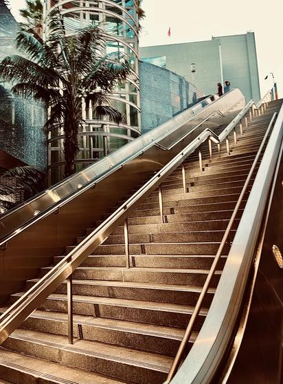 A color, low-angle, upward-facing photograph in portrait orientation that captures the outdoor concrete staircase and adjacent escalator at the Vermont/Sunset station in East Hollywood. The wide, multi-tiered stairs feature sleek, metallic handrails that create strong diagonal lines leading the eye toward the upper level. To the left, a vibrant green palm tree stands in front of a modern architectural structure with curved glass windows and blue-tiled walls. The warm, slightly sepia-toned lighting casts soft shadows across the stone steps, emphasizing the urban, geometric design of the transit hub. In the background, the station’s contemporary facade rises against a pale sky. A high-walled escalator bank runs parallel to the stairs, with the blurred figures of two people visible near the top, providing a sense of scale to the massive infrastructure. The metallic surfaces of the handrails and architectural accents reflect the ambient light, giving the scene a polished, metallic sheen. The combination of the tropical palm foliage and the sharp, industrial lines of the glass and steel architecture epitomizes the distinct urban aesthetic of Los Angeles public transit.