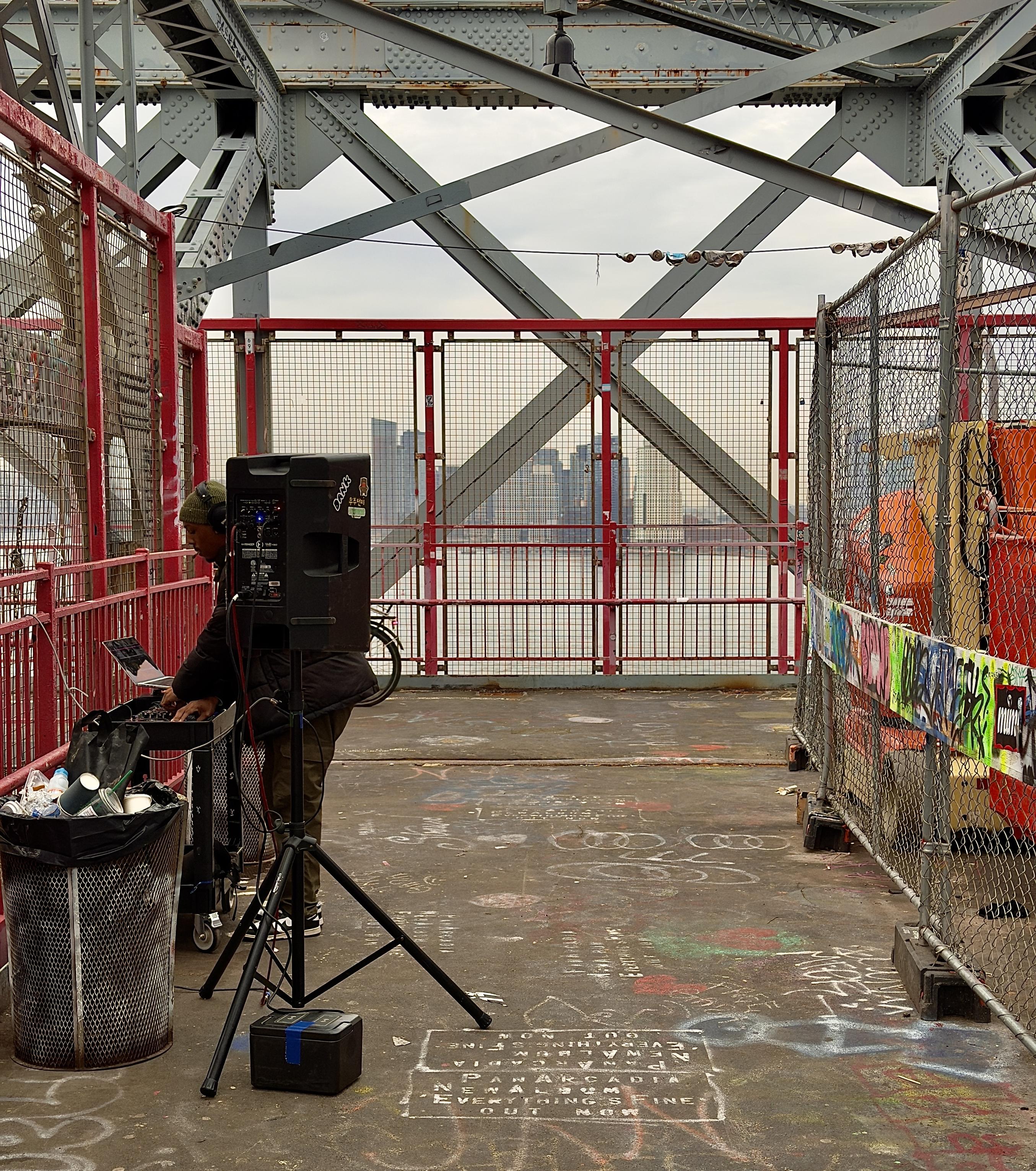 A color photograph in square aspect ratio. The focal point of the image is a person with dark skin standing on the pedestrian walkway of the Williamsburg Bridge, focused on a DJ setup. They wear a dark beanie, headphones, and a black puffer jacket over olive-colored pants. Their equipment includes a silver MacBook Pro perched atop a rolling black utility cart, which also holds a mixing console and various cables. To their right, a large black professional PA speaker stands tall on a tripod. In the foreground, a mesh trash can is filled full with discarded coffee cups and bottles visible, and the concrete ground is covered in white chalk graffiti and advertisements, one of which repeatedly mentions an album titled "Everything's Fine." The scene is framed by the heavy, industrial steel lattice of the bridge's superstructure, painted in a faded blue-grey, and red-painted safety fencing. Behind the DJ, the walkway opens up to a view of the East River and the hazy skyline of Manhattan in the distance under an overcast sky. To the right, a temporary construction area is cordoned off with chain-link fencing and orange equipment, partially obscured by a colorful banner covered in vibrant street art and stickers. The overall atmosphere is gritty and urban, capturing a spontaneous musical moment against a classic New York City backdrop.