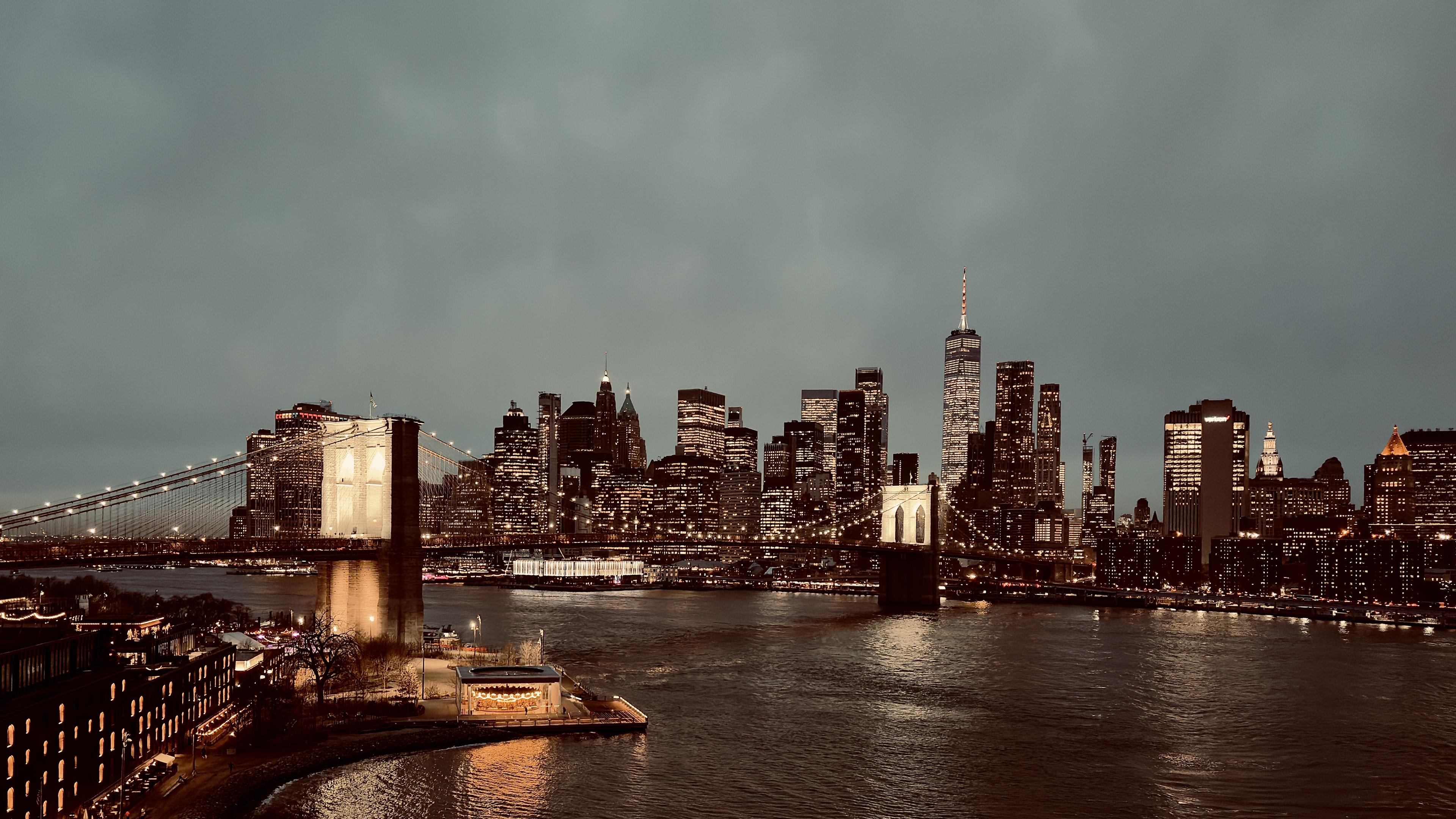 A color, wide-angle photograph taken at nighttime that captures the illuminated Brooklyn Bridge spanning across the East River, connecting to the dense, glowing skyline of Lower Manhattan. The bridge is the focal point, with its massive stone towers lit in a warm gold hue and its suspension cables draped like delicate strings of light across the dark water. In the foreground on the Brooklyn side, a brightly lit glass-enclosed carousel sits on the water’s edge, casting a vibrant reflection onto the river. The sky above is a heavy, overcast deep gray, which contrasts with the sparkling amber and white lights of the city. The Lower Manhattan skyline dominates the background with a silhouette of towering skyscrapers, featuring the prominent spire of One World Trade Center rising above the rest. Thousands of tiny lights from office windows and streetlamps create a shimmering texture against the dark buildings. The East River in the center of the frame is dark and slightly choppy, rippling with the mirrored glows of the city lights. The overall mood is atmospheric and serene.