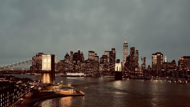 A color, wide-angle photograph taken at nighttime that captures the illuminated Brooklyn Bridge spanning across the East River, connecting to the dense, glowing skyline of Lower Manhattan. The bridge is the focal point, with its massive stone towers lit in a warm gold hue and its suspension cables draped like delicate strings of light across the dark water. In the foreground on the Brooklyn side, a brightly lit glass-enclosed carousel sits on the water’s edge, casting a vibrant reflection onto the river. The sky above is a heavy, overcast deep gray, which contrasts with the sparkling amber and white lights of the city. The Lower Manhattan skyline dominates the background with a silhouette of towering skyscrapers, featuring the prominent spire of One World Trade Center rising above the rest. Thousands of tiny lights from office windows and streetlamps create a shimmering texture against the dark buildings. The East River in the center of the frame is dark and slightly choppy, rippling with the mirrored glows of the city lights. The overall mood is atmospheric and serene.