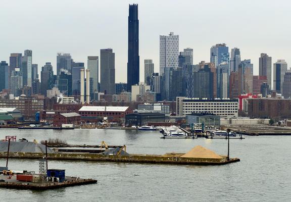 A color, wide-angle photograph taken from an elevated view of the Downtown Brooklyn skyline behind the industrial waterfront of the East River under a pale, overcast afternoon sky. In the foreground, a concrete pier extends into the choppy grey water, featuring a large mound of sand, yellow excavators, and various construction equipment. Further back, the Brooklyn Navy Yard area displays low-slung brick warehouses and a pier where several white commuter ferries are docked. Two prominent white smokestacks stand out against the mid-ground industrial buildings, adding a vertical element to the lower half of the composition. The background is dominated by a dense cluster of modern skyscrapers in varying shades of brown, grey, blue, and white. The most striking feature is the Brooklyn Tower, a very tall, slender, dark-bronze-to-black skyscraper with a distinctive neo-Gothic, fluted texture that tapers toward the top. Surrounding it are several glass-faced residential and office towers. The overall color palette is muted, consisting primarily of industrial greys, earthy brick tones, and the deep metallic dark of the central tower.