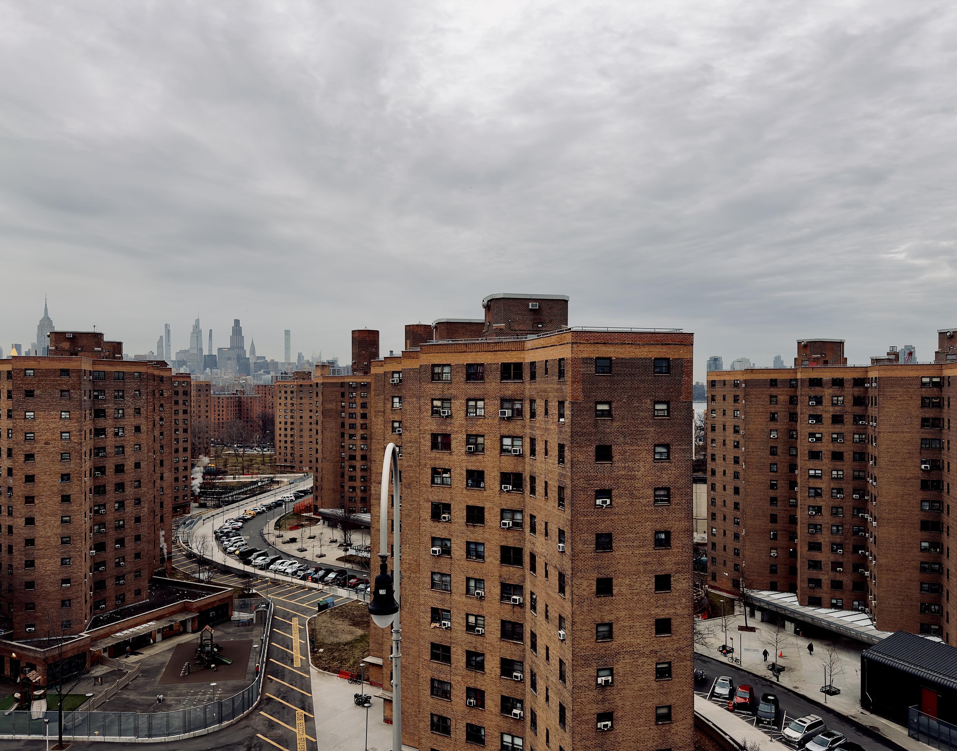 A color photograph in square aspect ratio. An elevated, high-angle view looks north over a dense complex of multi-story, red-brick housing buildings in the Lower East Side neighborhood of Manhattan. The foreground and middle ground are dominated by several identical, cruciform-shaped apartment towers with many small windows and protruding air conditioning units. Between the buildings, a curved asphalt road is lined with parked cars, and a small playground with a green slide is visible at the bottom left. The sky is a heavy, uniform grey with thick cloud cover, casting a flat, somber light over the urban landscape. In the far distance, the midtown Manhattan skyline rises as a jagged silhouette against the overcast horizon. Iconic skyscrapers, including the Empire State Building and several slender "pencil towers," are visible, though somewhat hazy due to the distance and weather conditions. A glimpse of the East River is visible to the right between the apartment blocks.
